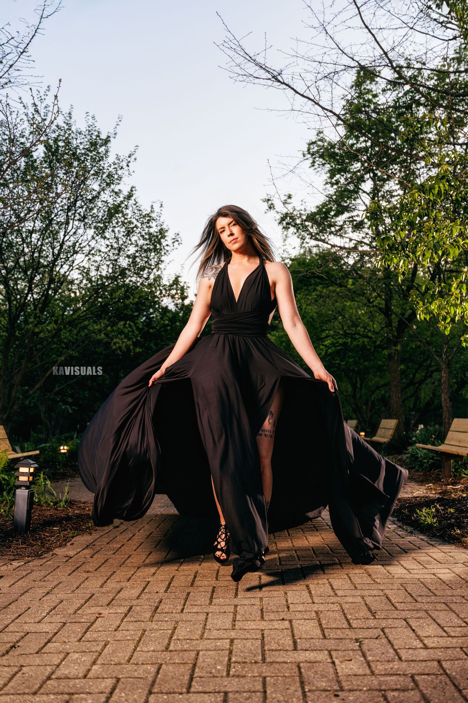 Woman in black flowing dress twirling on brick pathway at dusk.