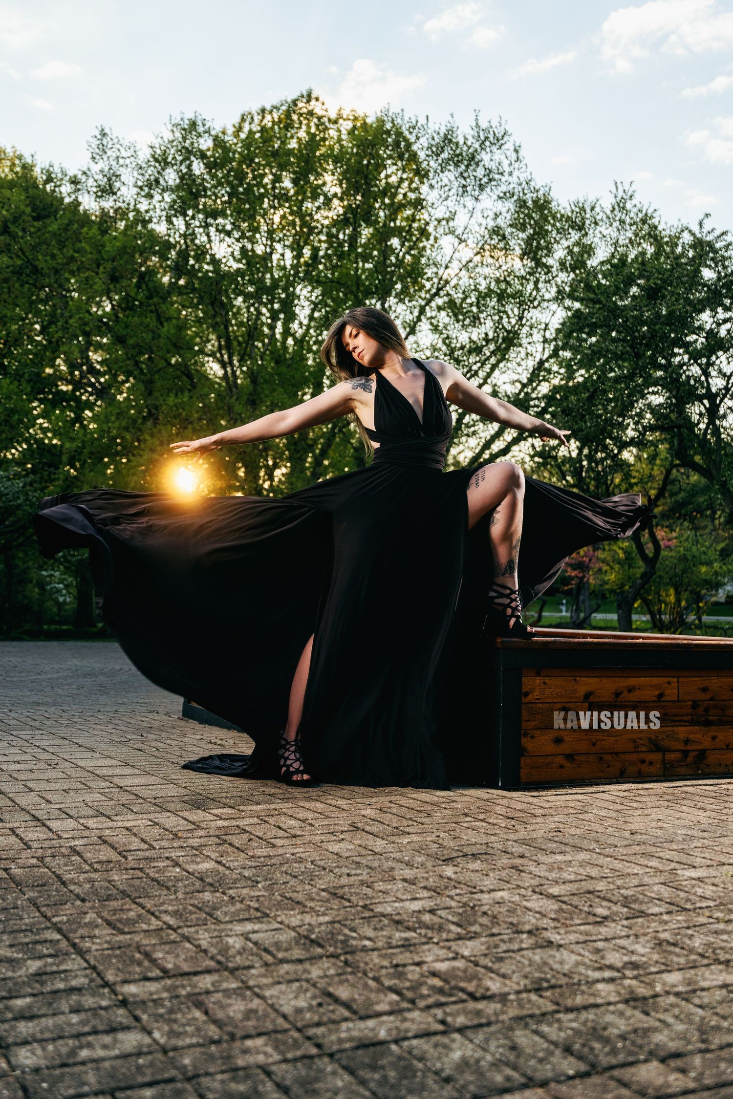 Woman in black dress with flowing fabric standing on wooden platform at sunset.