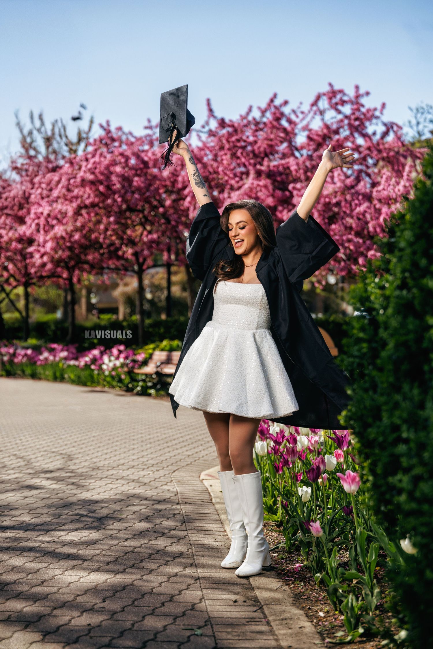 Woman in white dress and graduation gown celebrating outdoors near blooming pink cherry blossom trees.