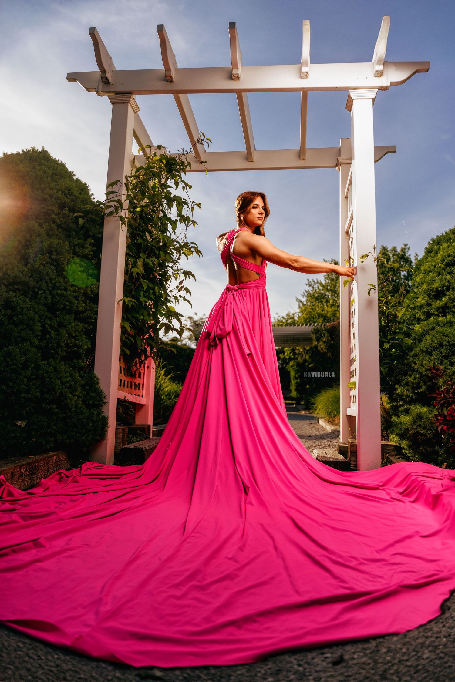 Woman in elegant pink gown with long train posing under white pergola arch.