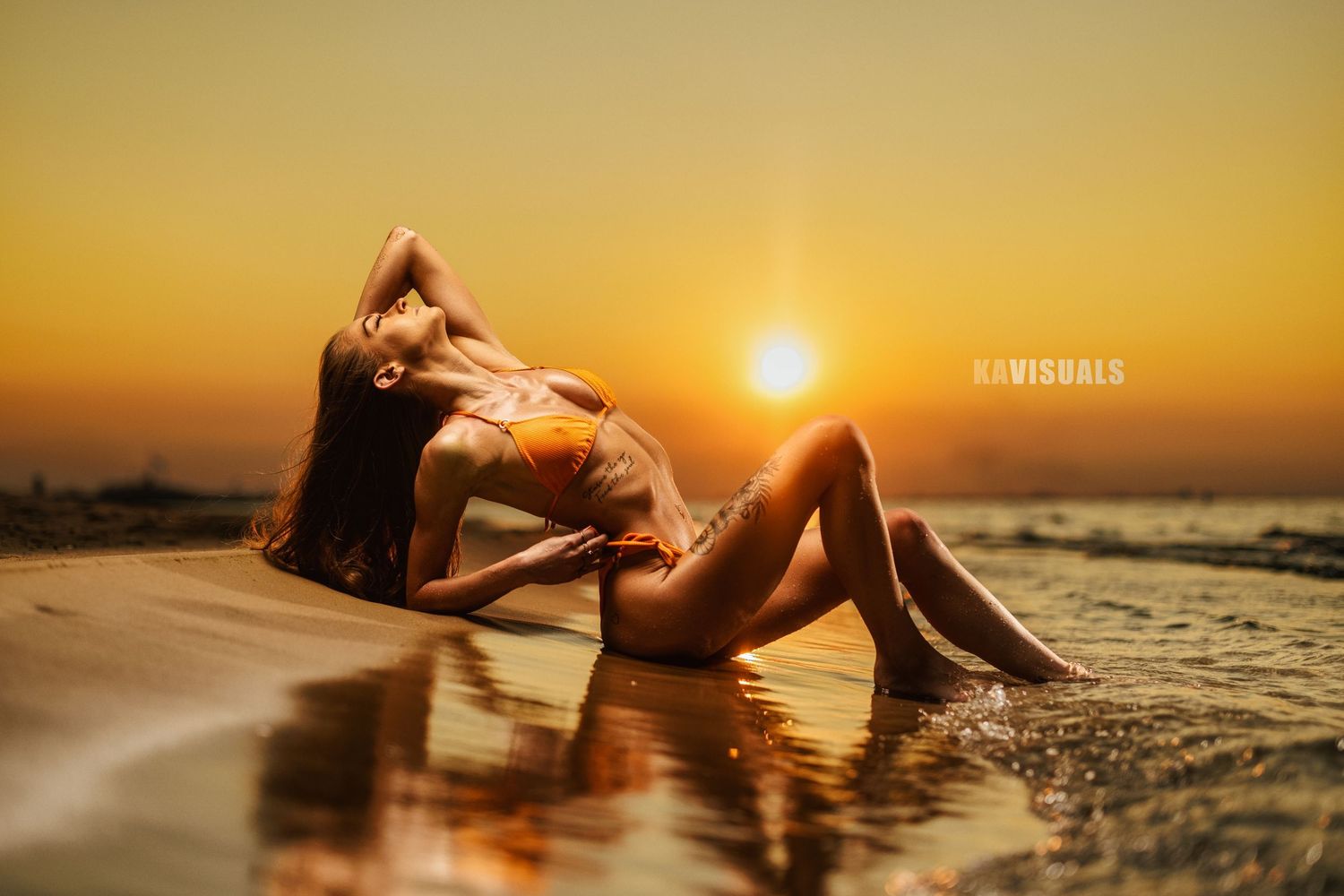 Woman in orange bikini posing on wet beach sand during golden sunset.