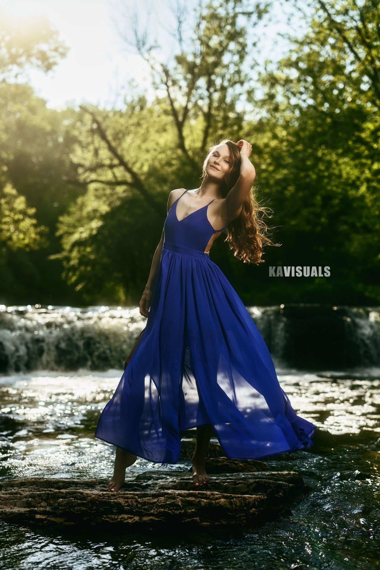 Woman in flowing royal blue dress stands on rocks near waterfall in sunlit forest.