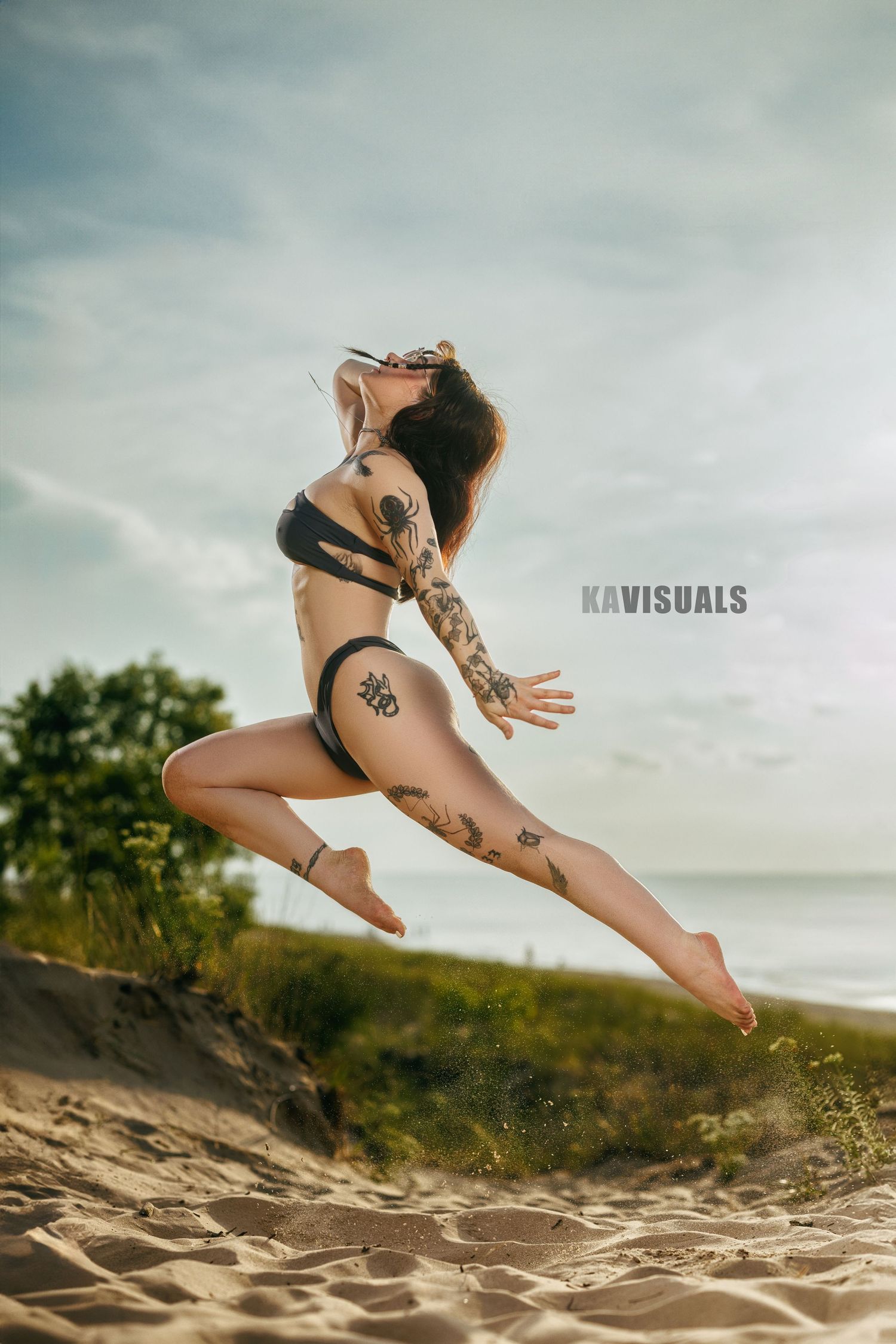 Tattooed woman in black bikini jumps on sandy beach under cloudy sky.