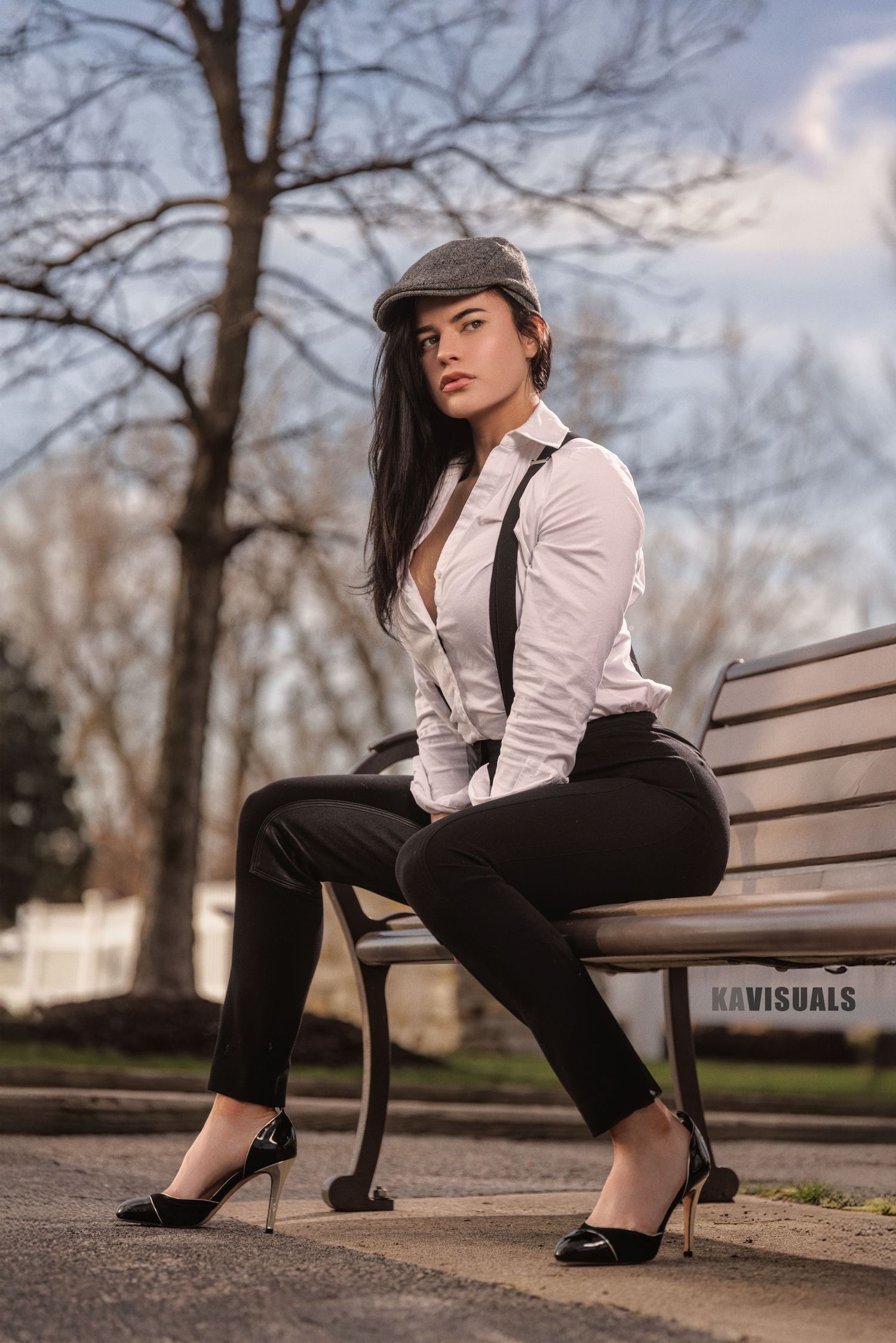 Elegant woman in white shirt, suspenders, and flat cap sitting on park bench.