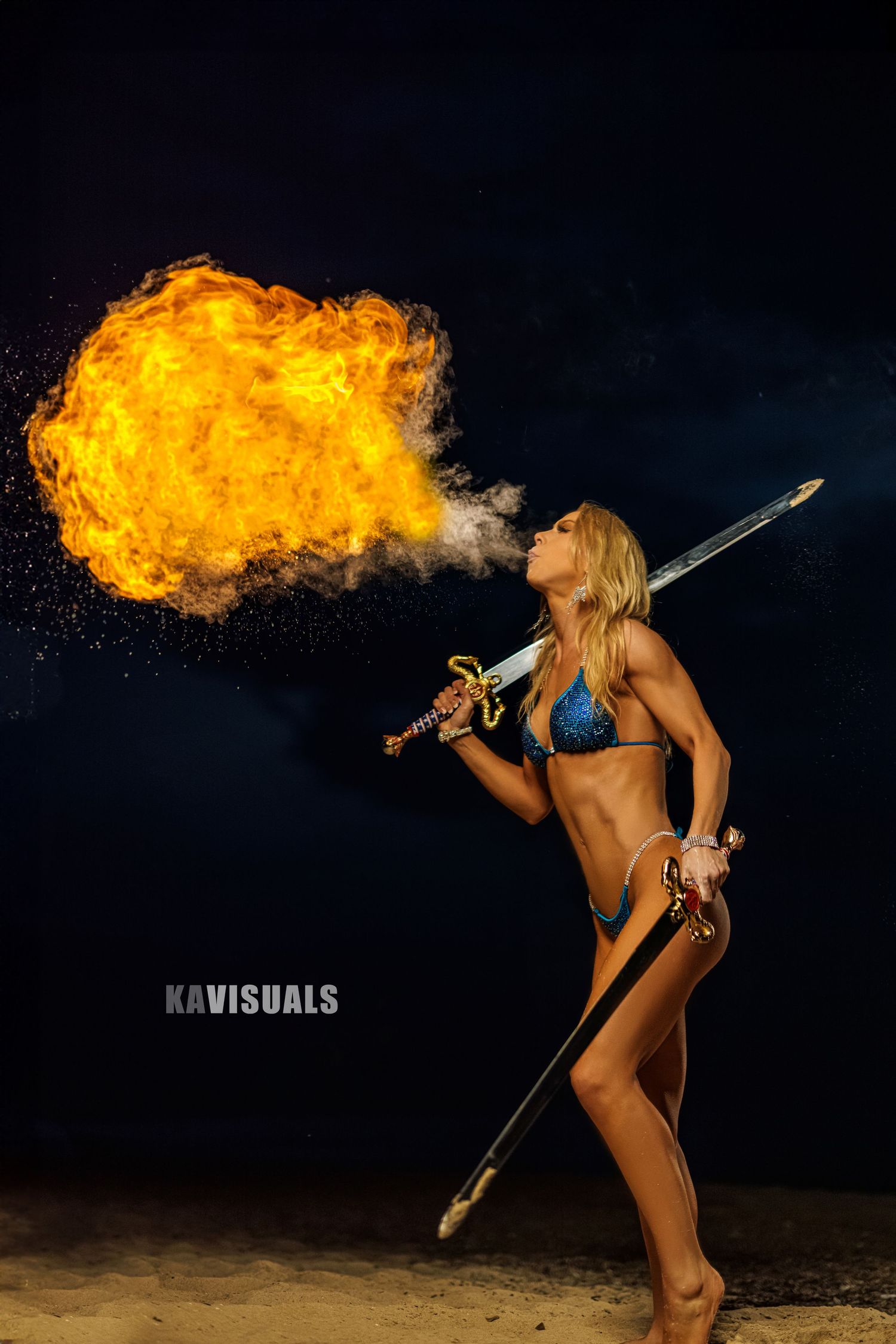 Athletic blonde woman breathes fire while holding a sword in a dramatic night shoot.