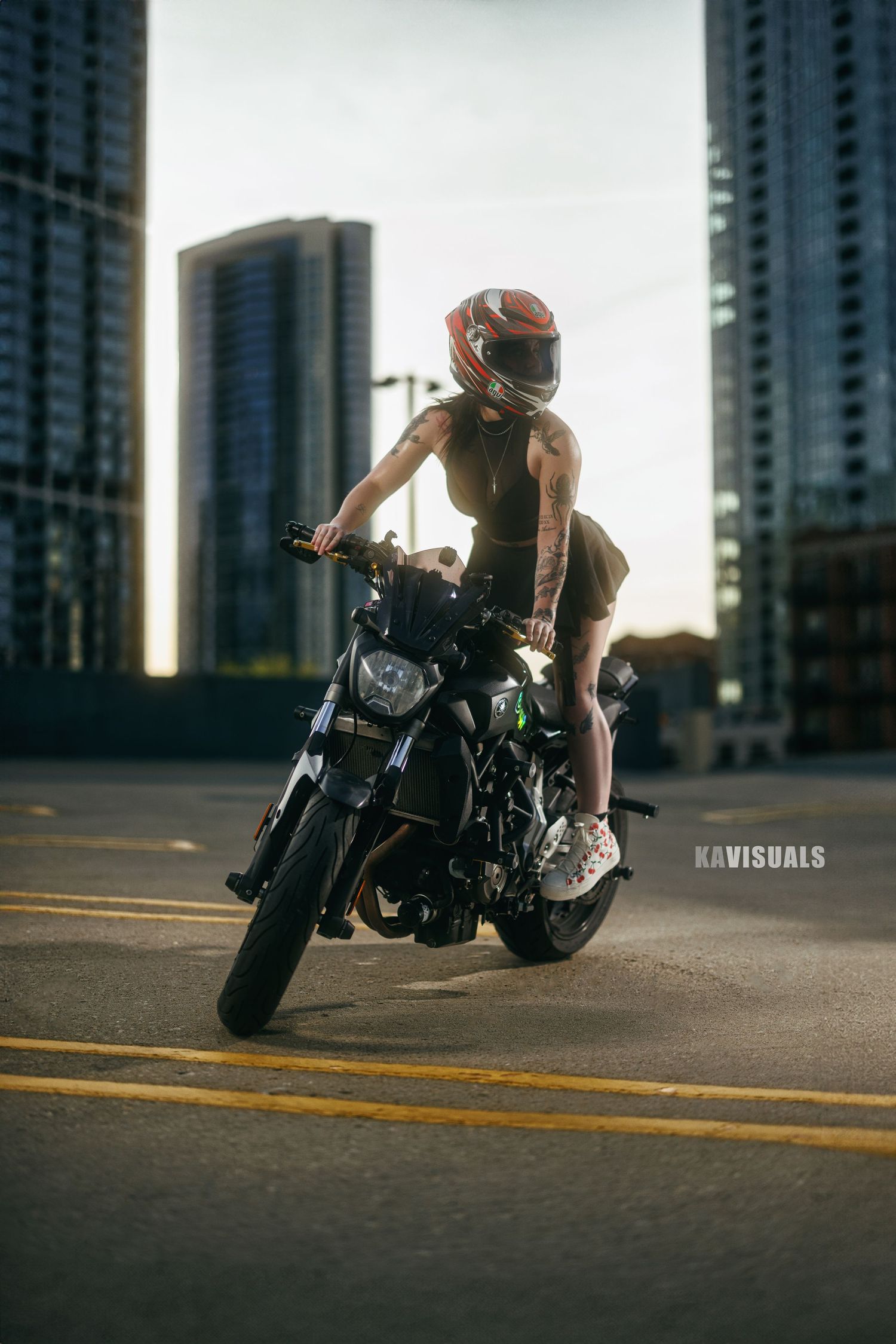 Female motorcyclist wearing red helmet riding a black motorcycle on an urban rooftop parking lot at dusk.