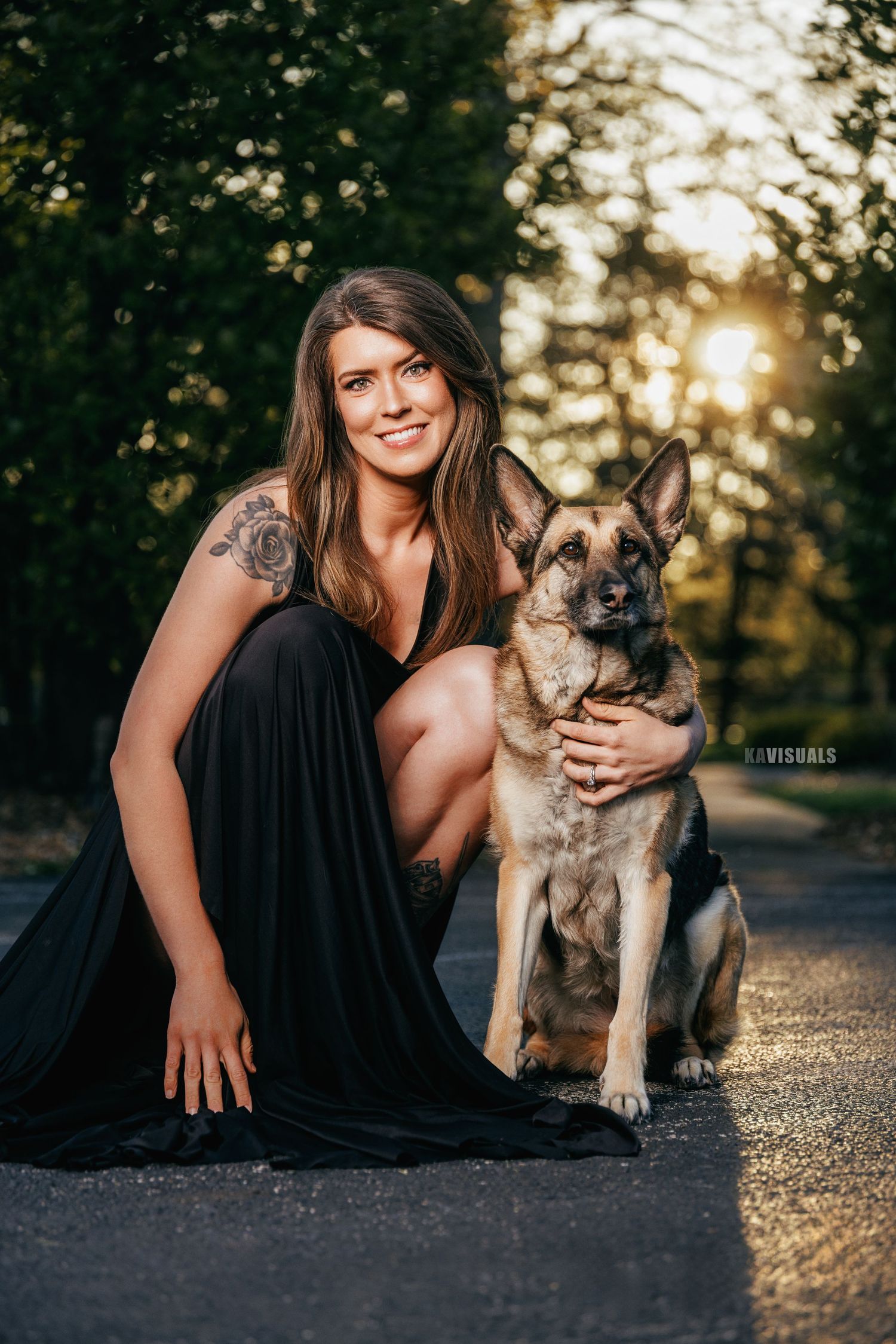 Woman in a black dress posing with a German Shepherd dog on a sunlit forest road.