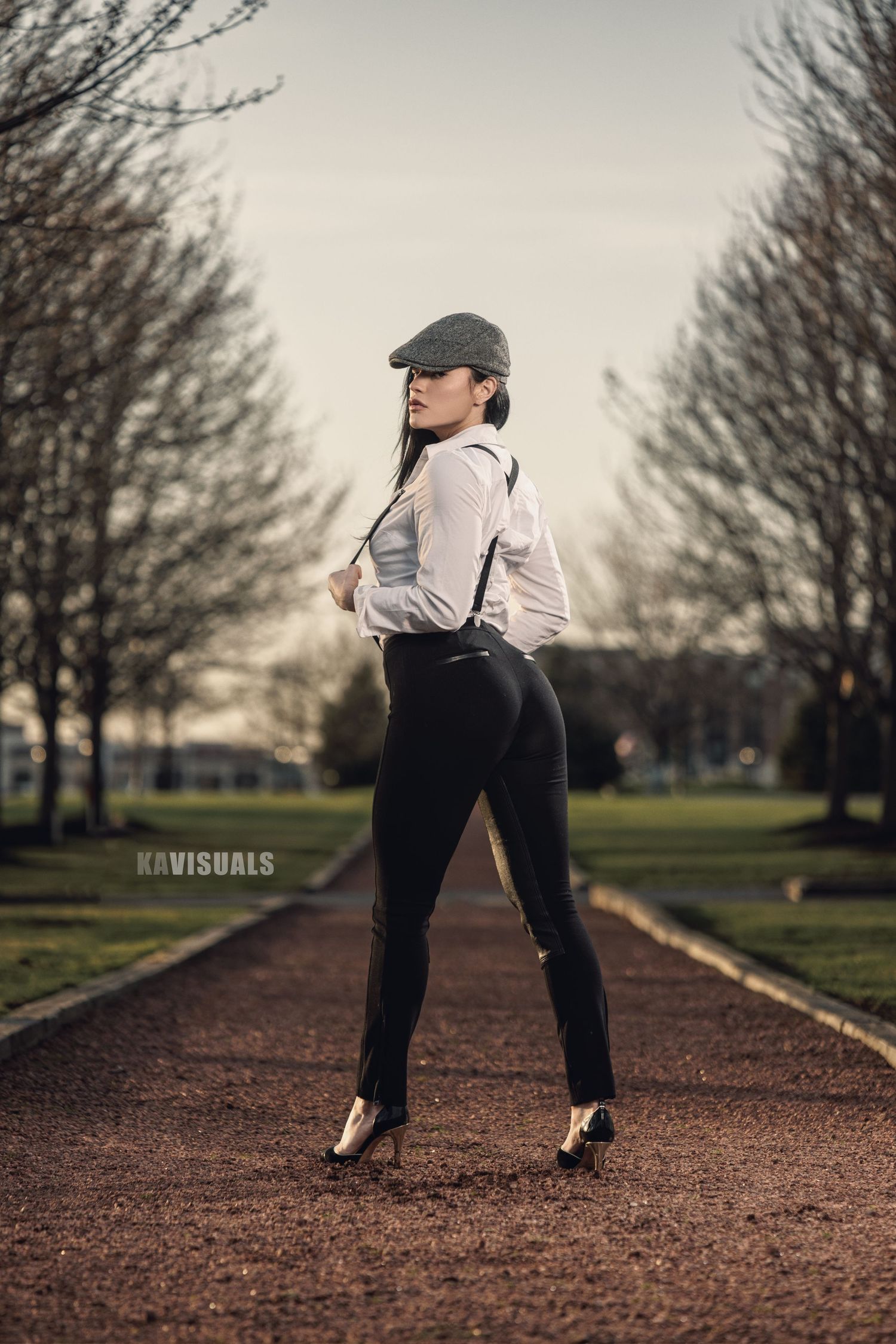 Stylish woman in white shirt, suspenders, and flat cap posing in park pathway.
