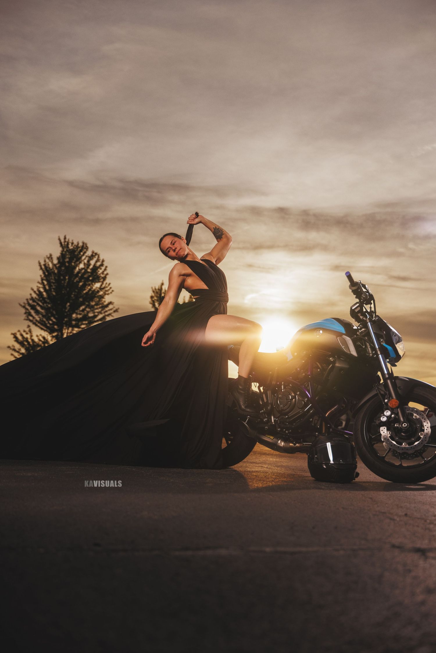 Woman in a flowing black dress posing dramatically beside a motorcycle at golden hour sunset.