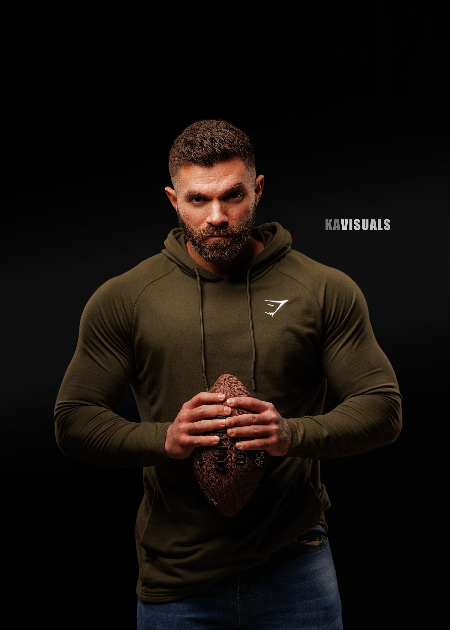 Muscular man in olive hoodie holds football against dark studio background.