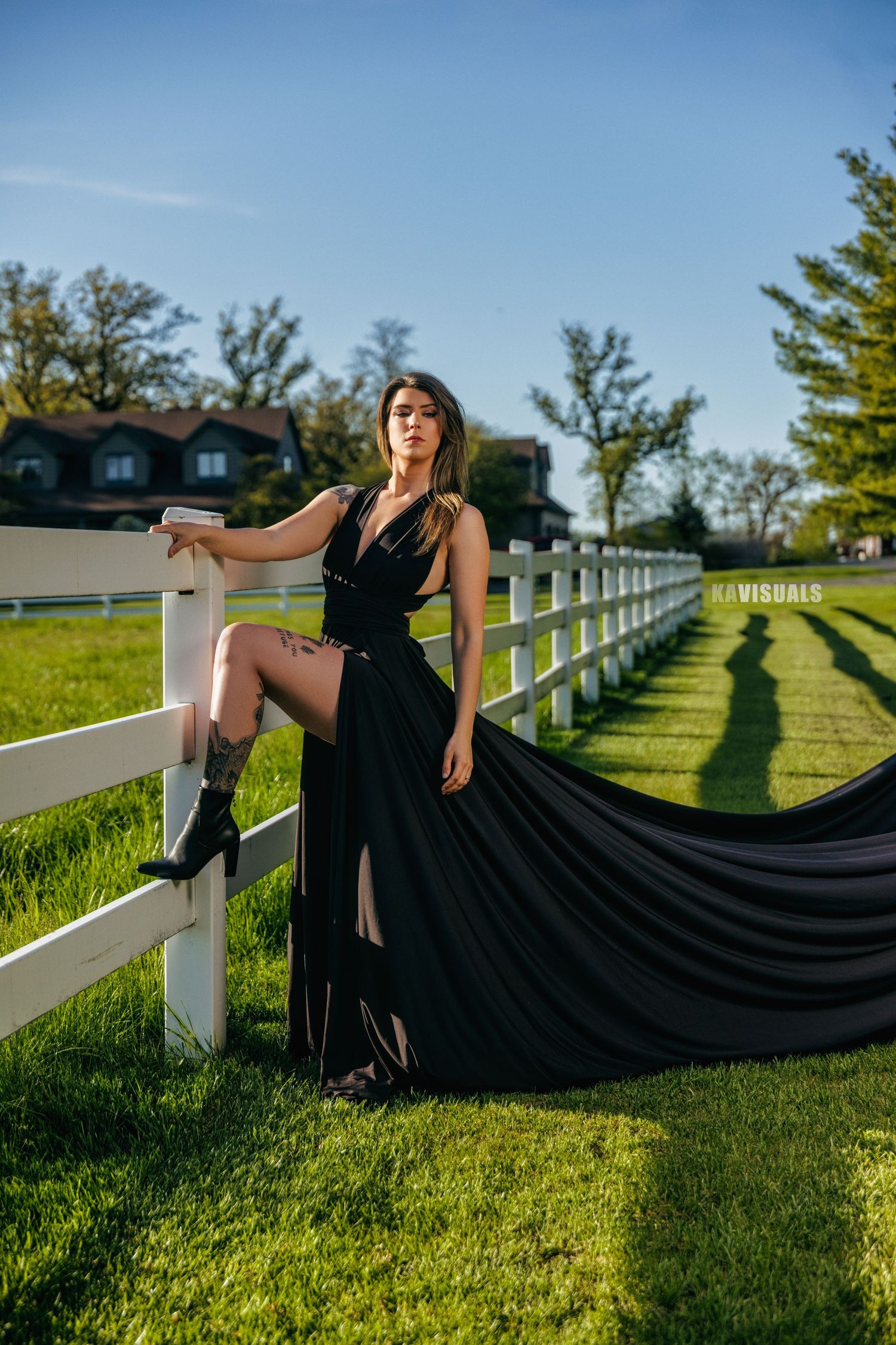 Elegant woman in flowing black gown posing by a white fence on a sunny green farm.