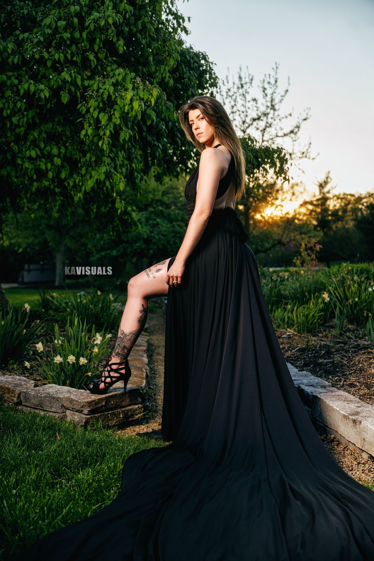 Woman in black gown with train posing in garden at golden hour.