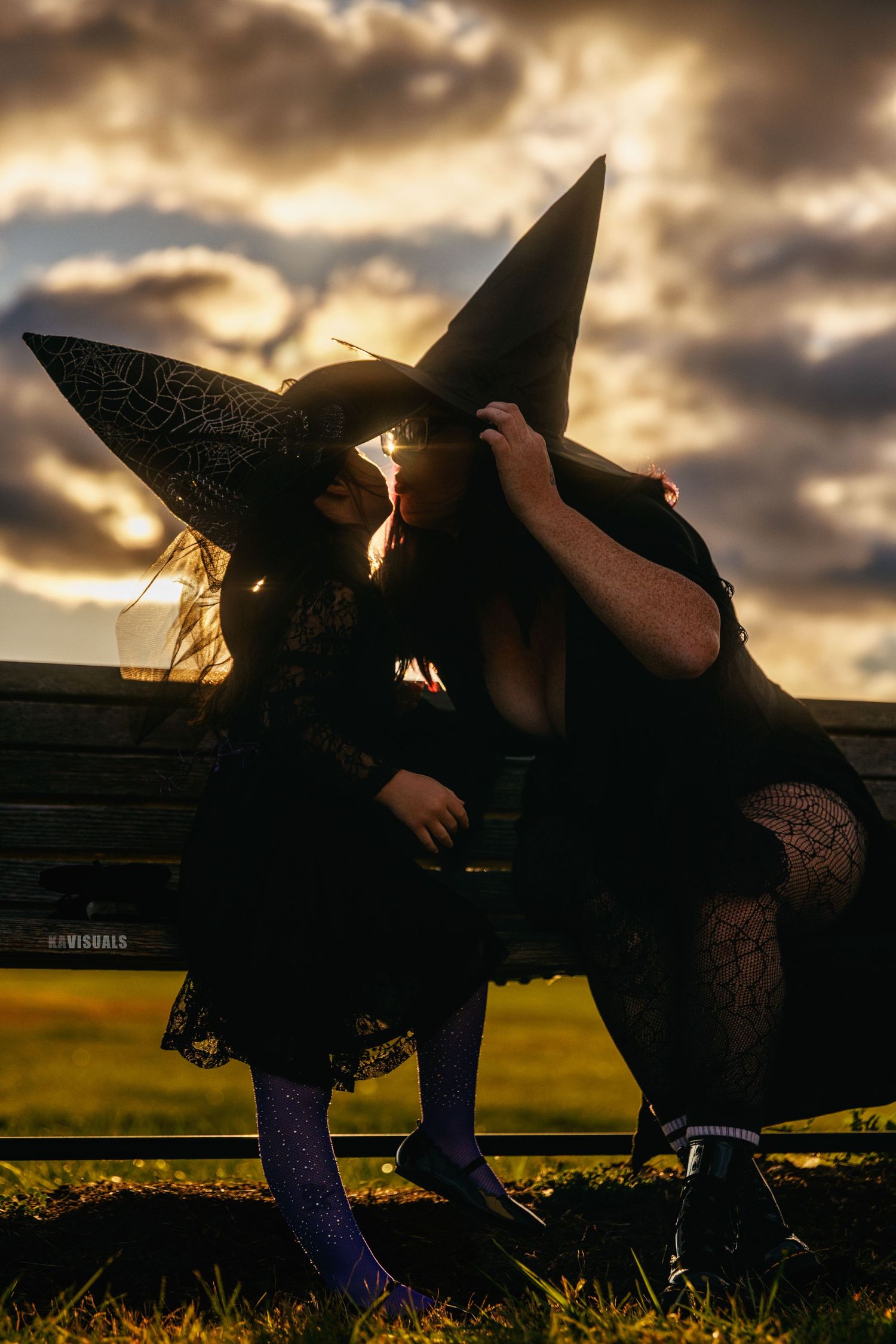 Two women in witch costumes kissing on bench at sunset during Halloween.