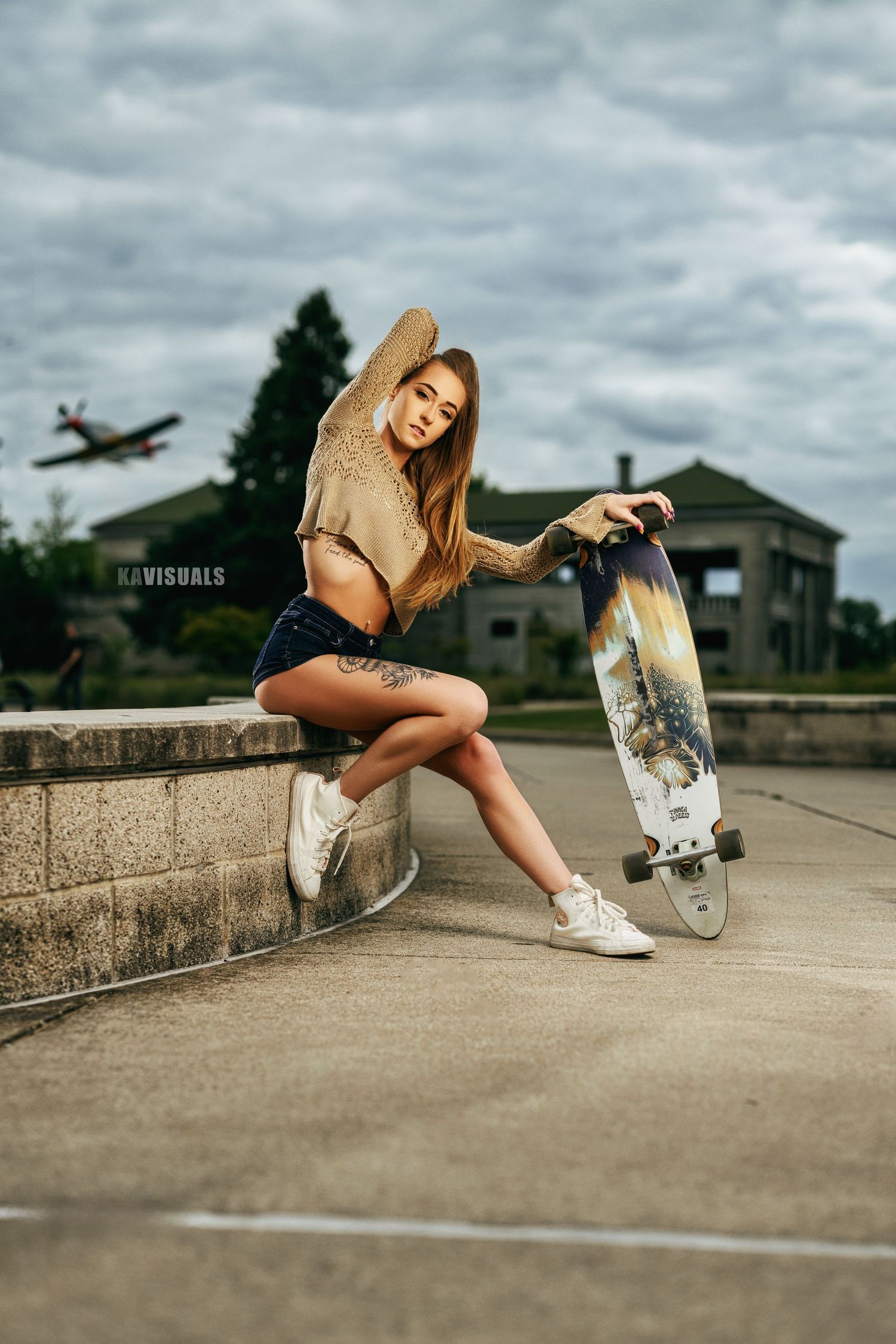 Tattooed blonde woman in denim shorts holding longboard skateboard while sitting on concrete ledge outdoors.