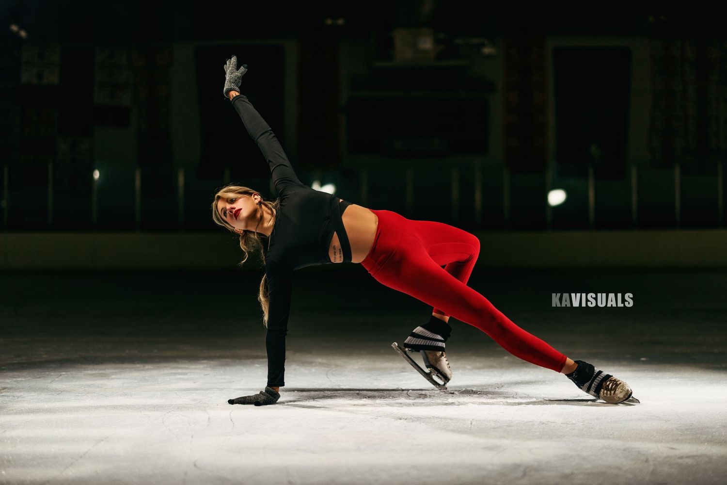 Female figure skater in red leggings performing a side plank balance pose on ice at an indoor rink.