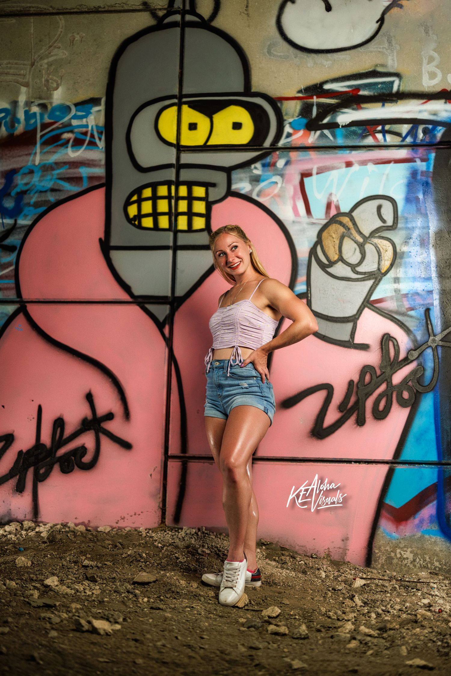Blonde woman in denim shorts posing before a colorful Bender graffiti mural wall.