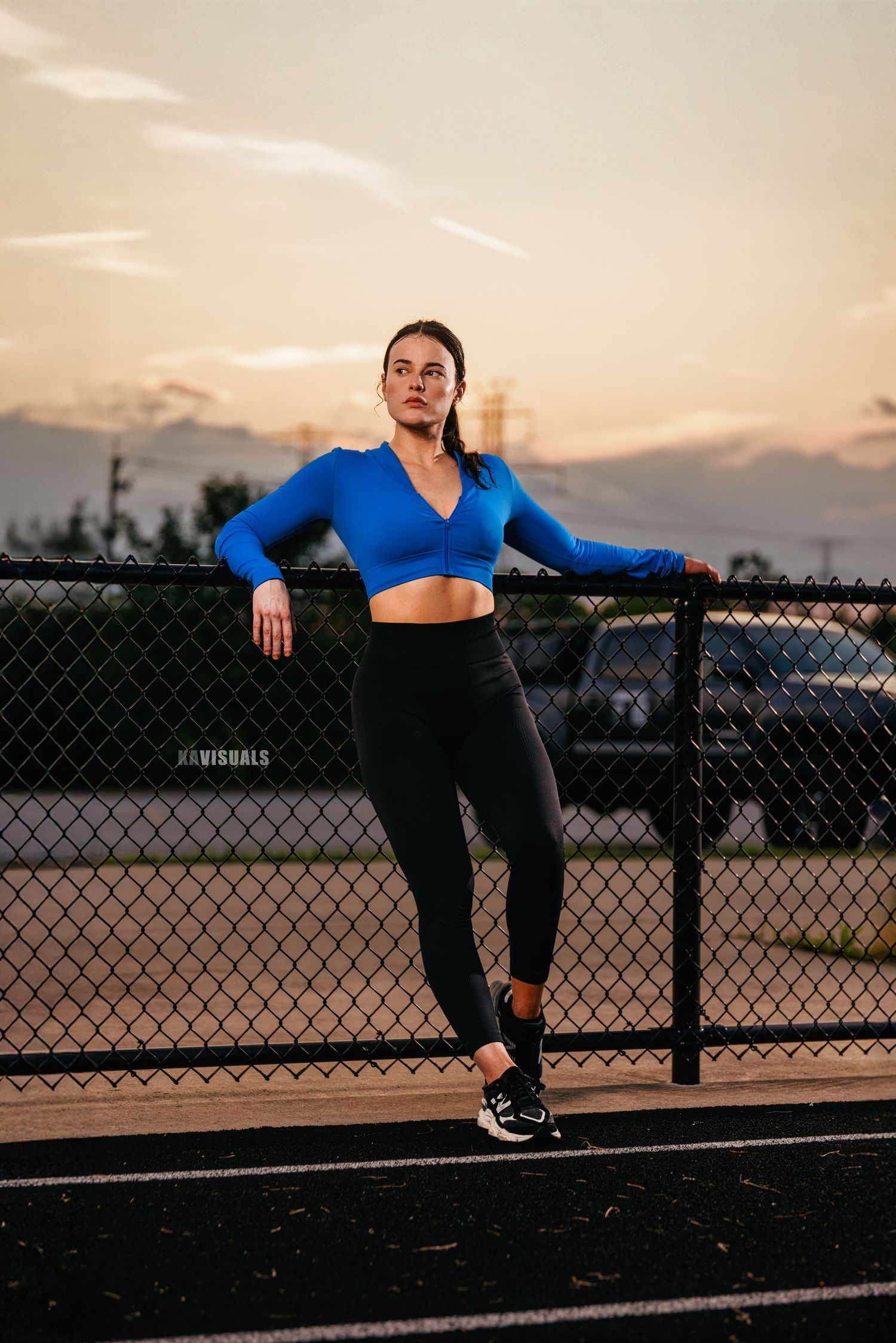 Fit woman in blue crop top and black leggings leaning on chain-link fence at sunset.