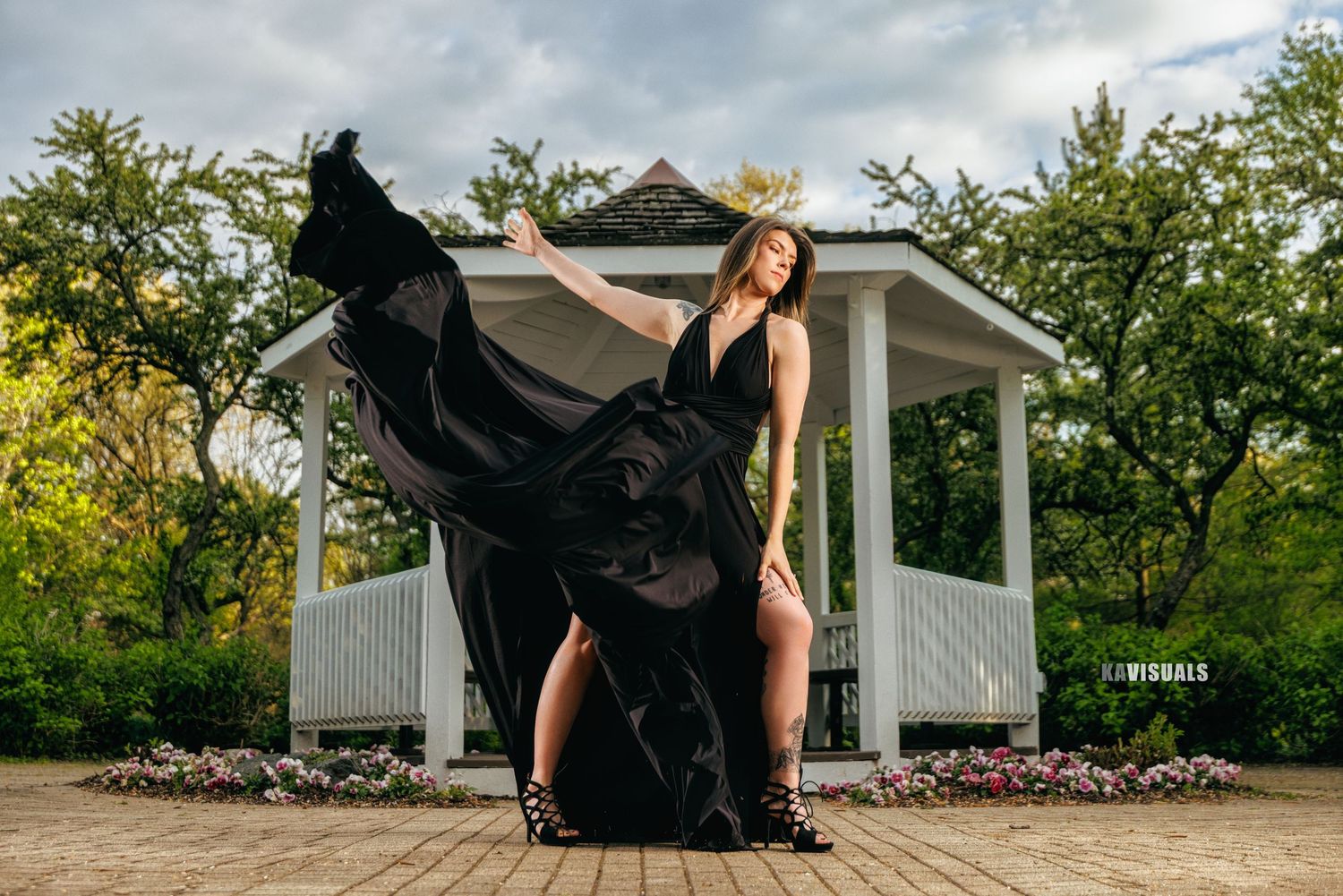 Woman in black flowing gown near white gazebo in park setting.