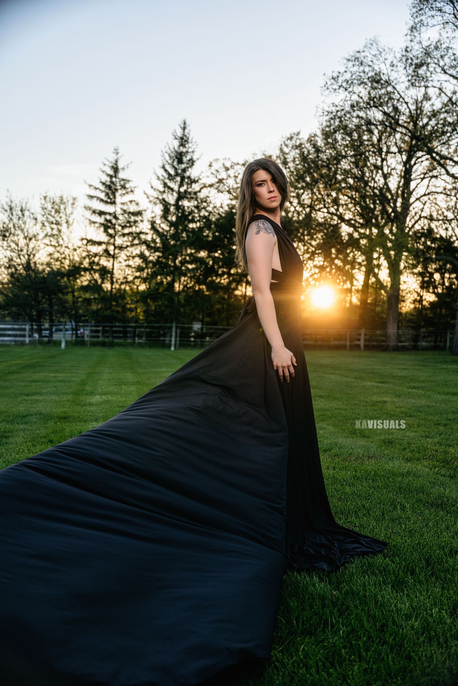 Woman in dramatic black gown with long train standing in a field at sunset.
