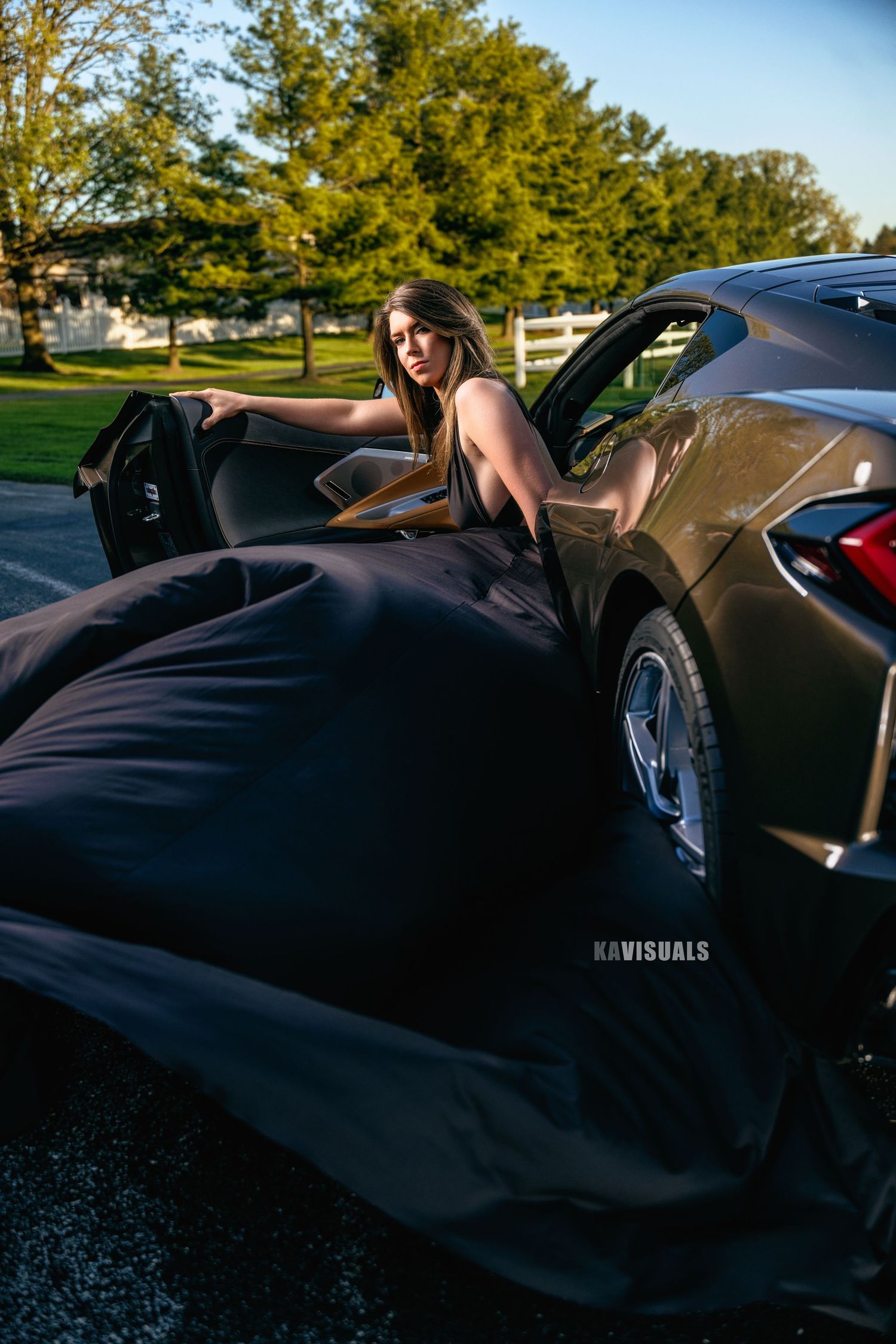 Woman in black flowing gown emerging from dark sports car door in sunlit outdoor parking area.