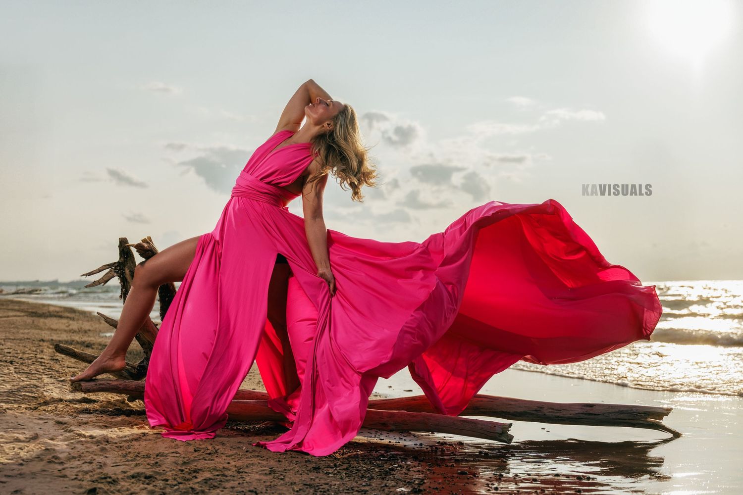 Woman in flowing pink dress dancing on a beach with fabric billowing in wind.