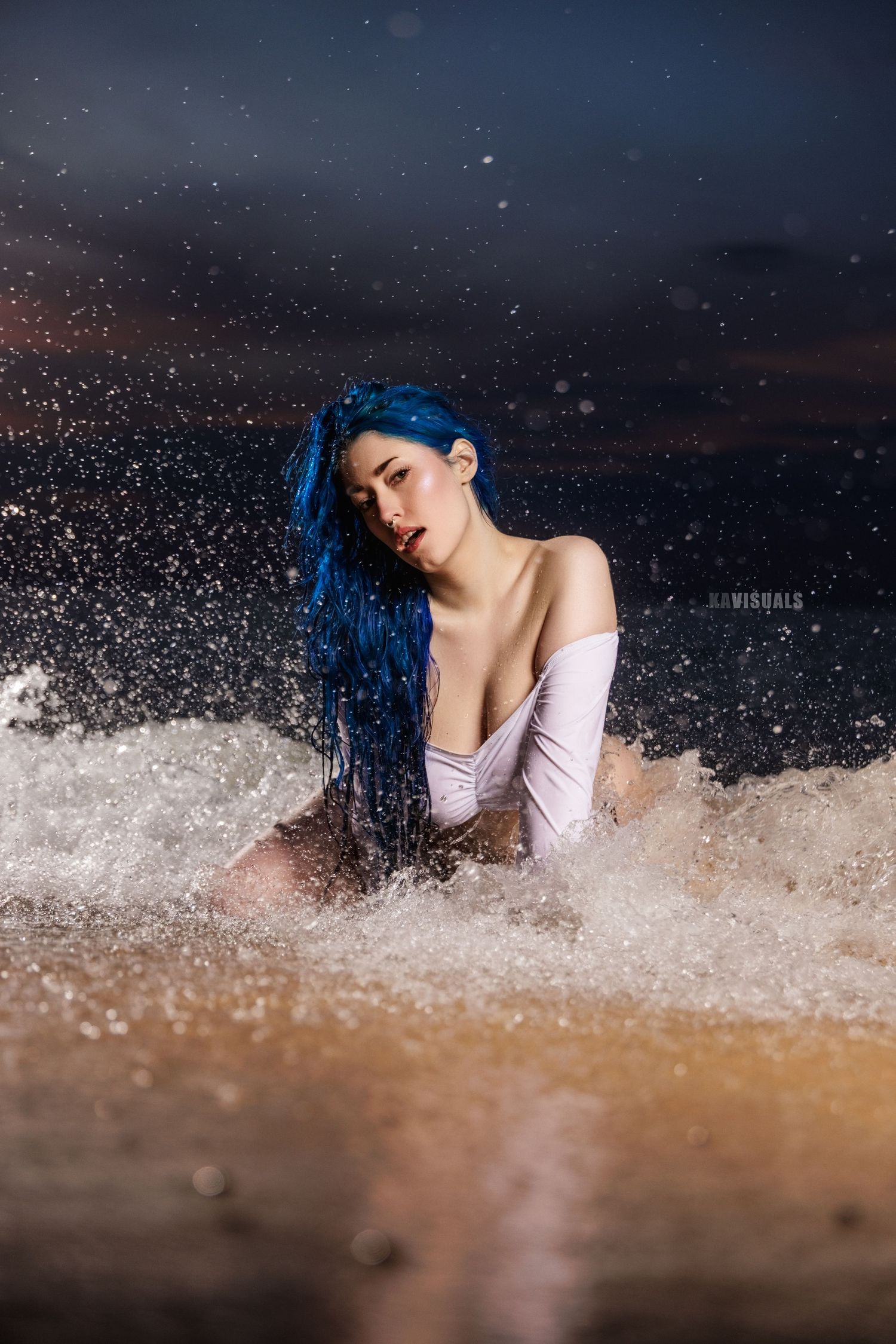 Woman with blue hair in white top sitting in ocean waves at night with dramatic water splashes.