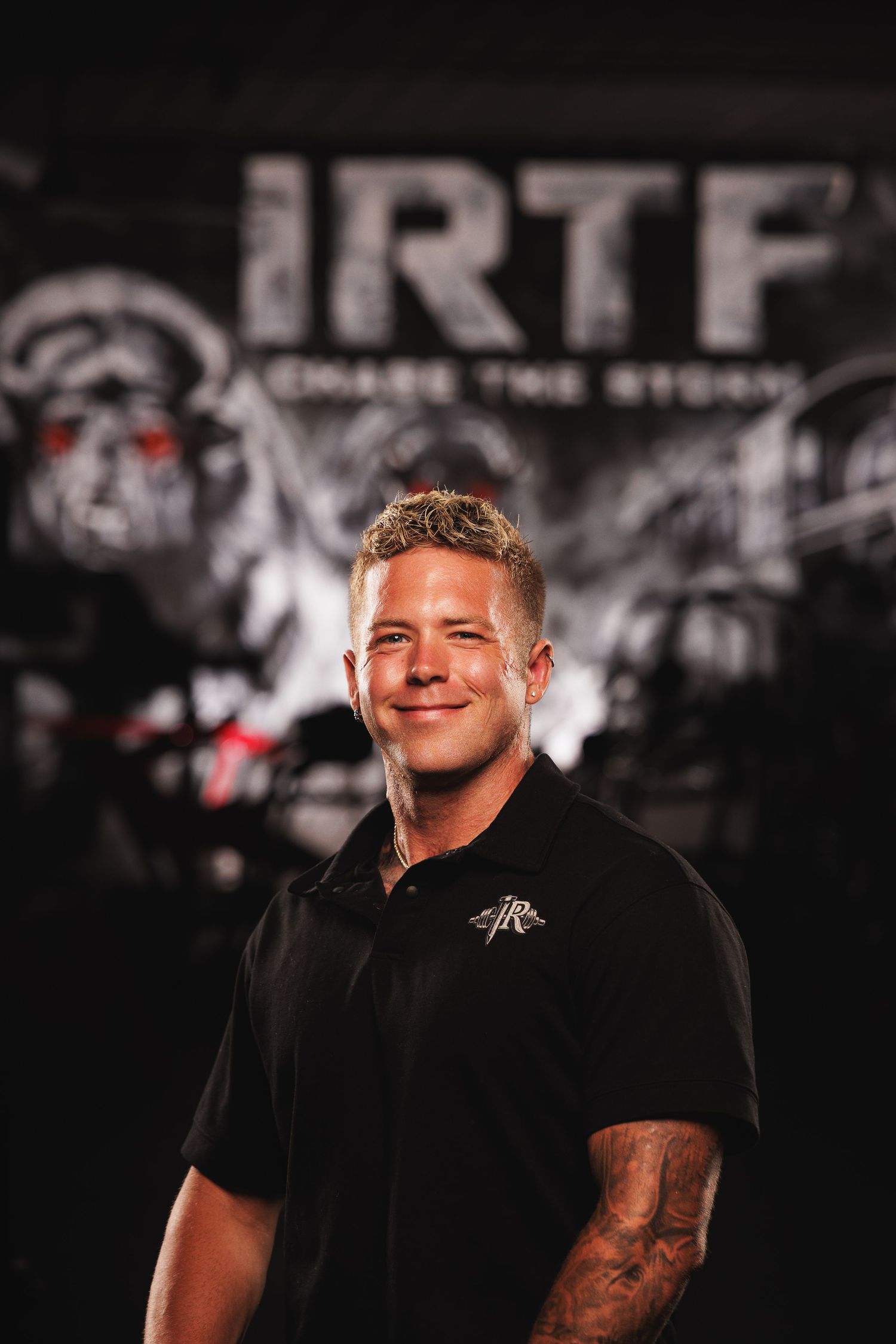Smiling man in black IRTF polo shirt posing before branded gym backdrop with red-eyed logo.