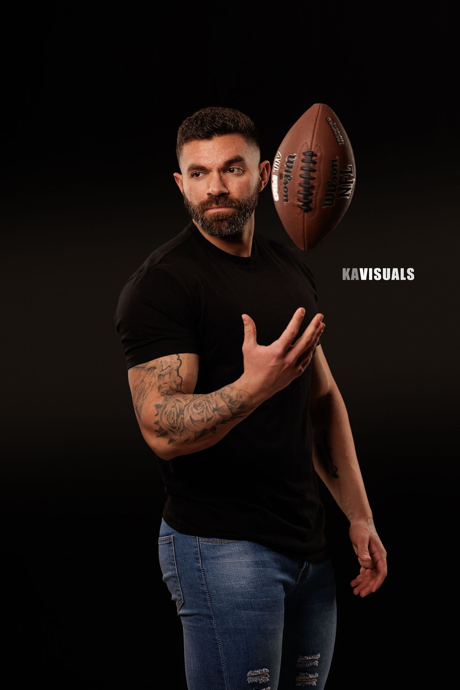 Tattooed bearded man in black tee tosses football on dark background.
