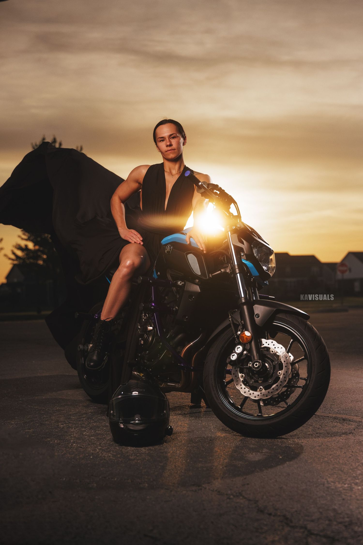 Woman in a black outfit leaning against a blue motorcycle with helmet on ground at sunset.