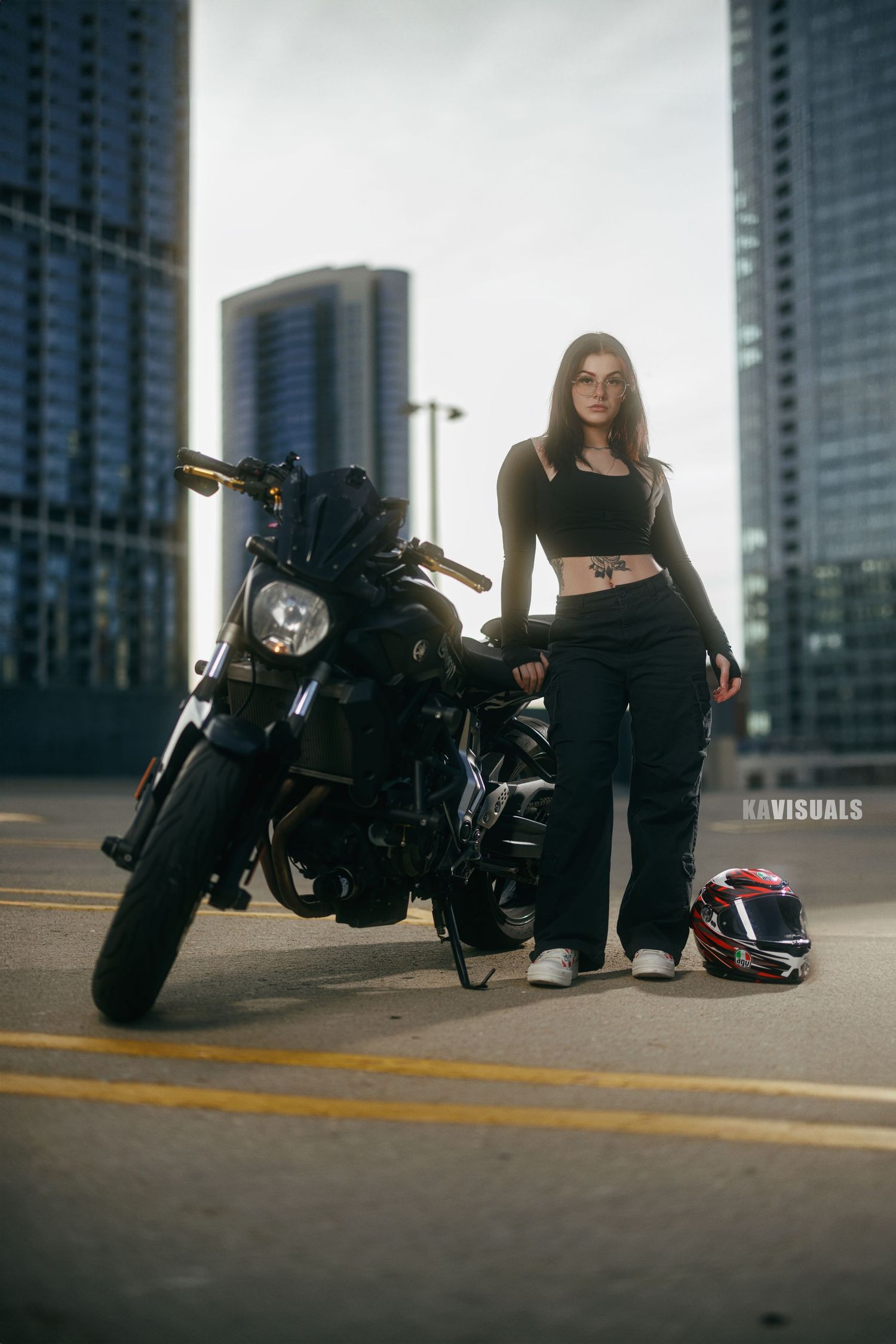 Young woman in black crop top and cargo pants standing beside a black motorcycle on a rooftop parking lot with city skyline.