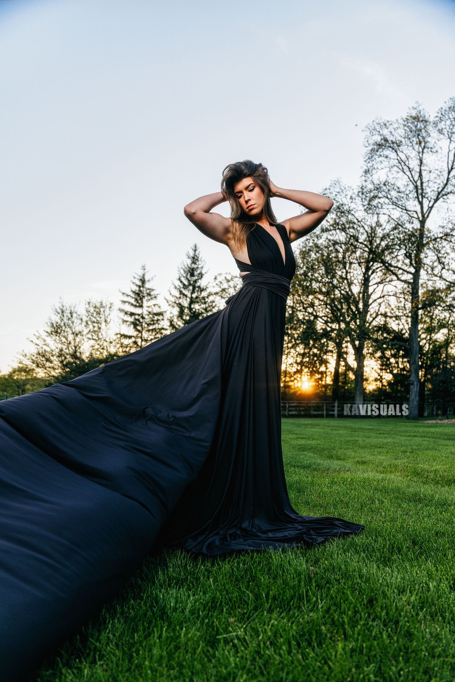 Woman in black gown with long train on green lawn at sunset.