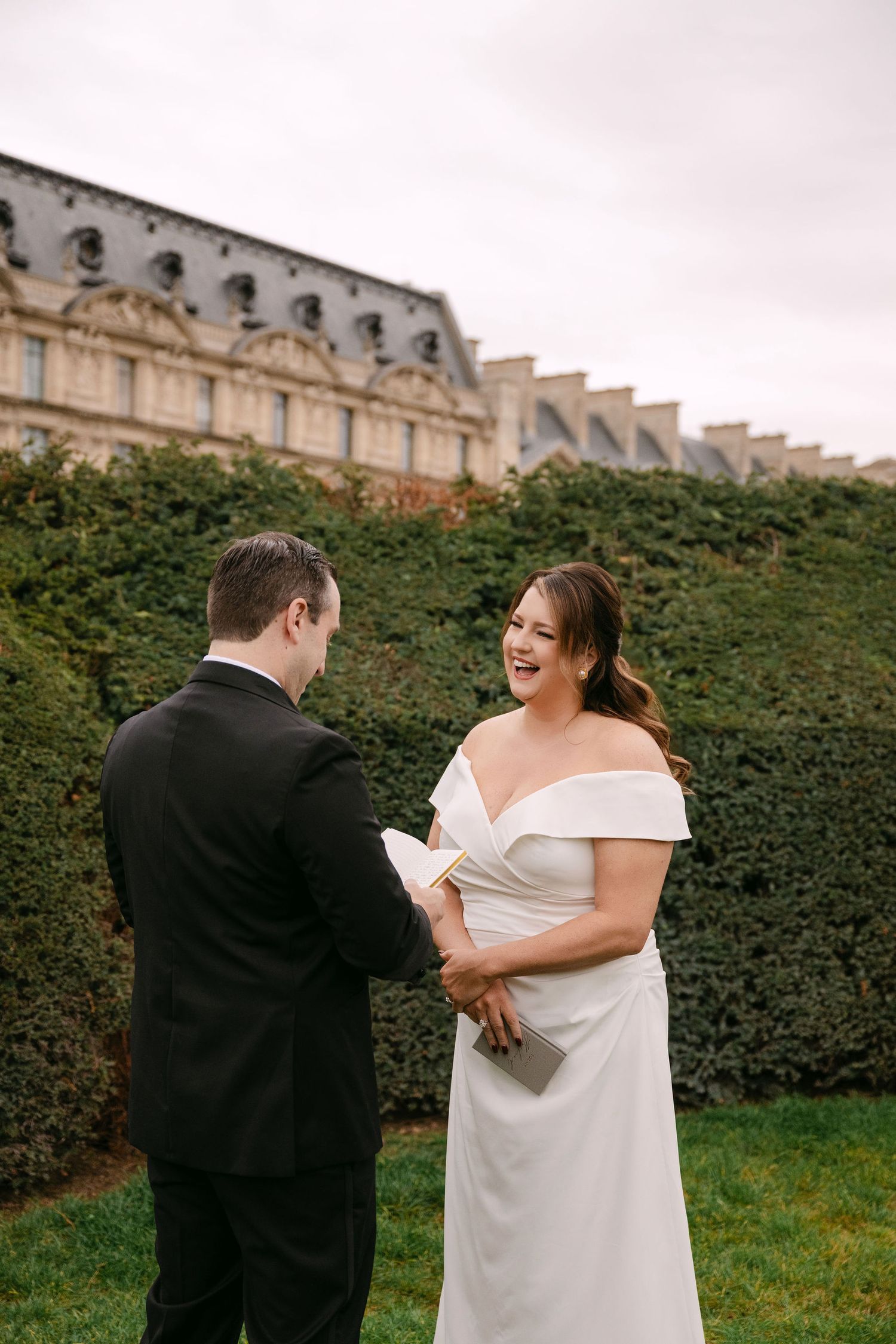 A couple shares a joyful moment together in front of a manicured hedge wall and historic building facade.
