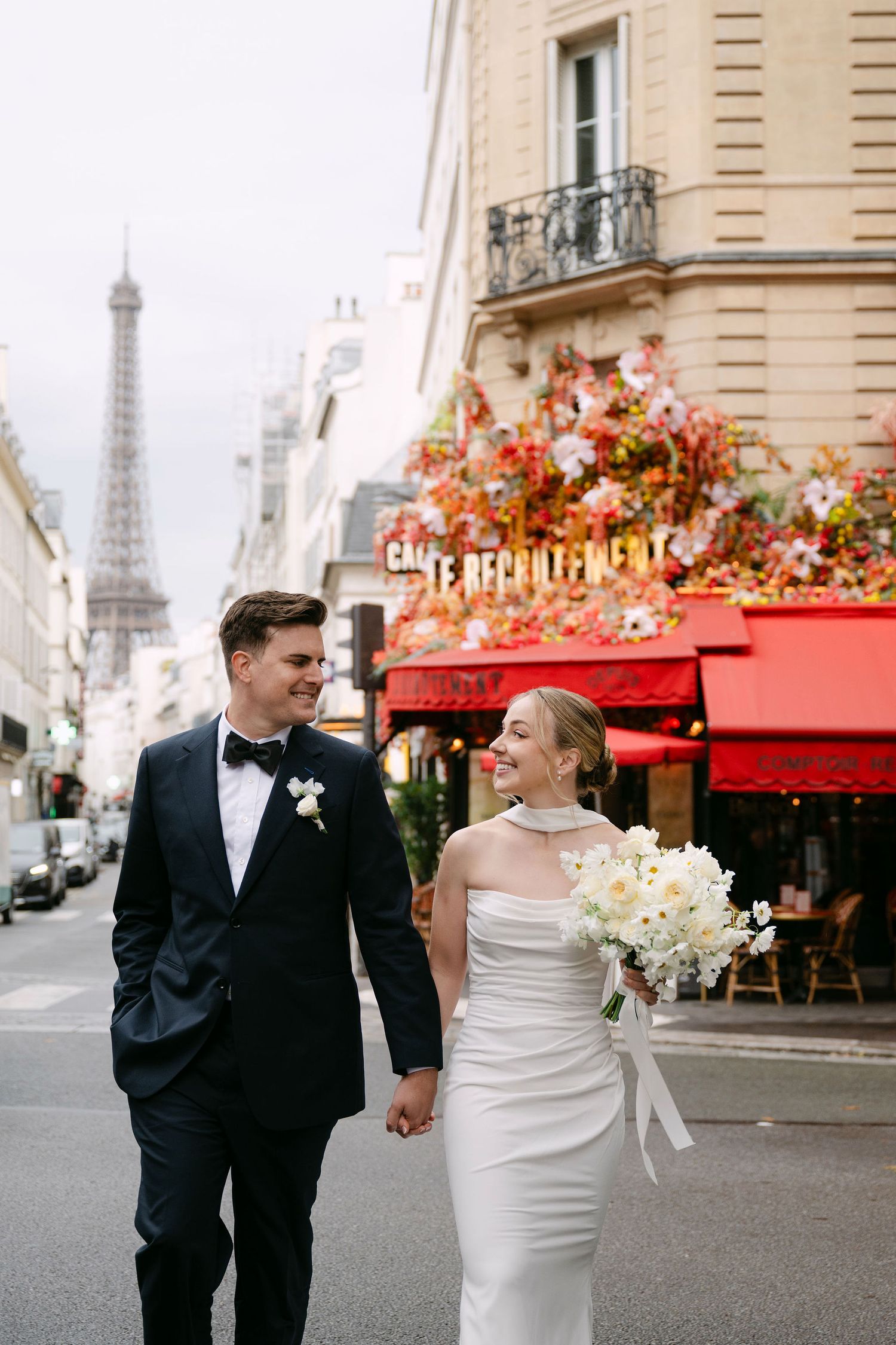 Newlywed couple walking along charming Paris street with Eiffel Tower and red cafe awnings in background.