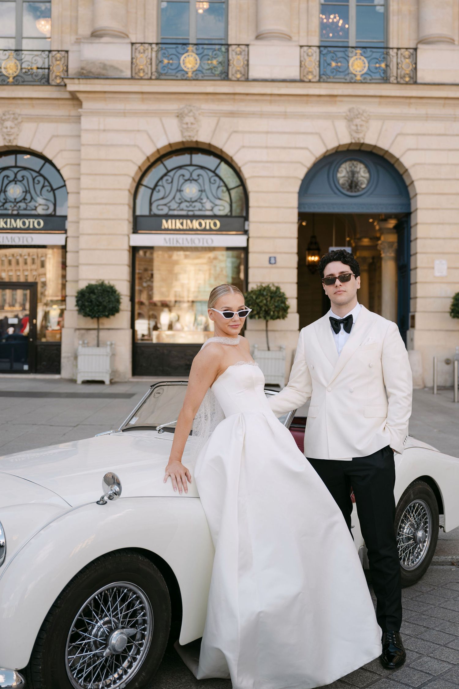 A bride and groom pose elegantly next to a classic vintage car outside a grand European building.
