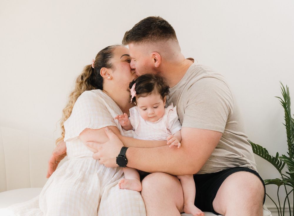 Parents kissing their baby girl on the cheeks during a bright, minimalist family portrait session.
