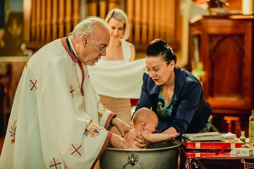 Priest performing Catholic baptism ceremony, pouring water over infant held by mother over baptismal font in church.