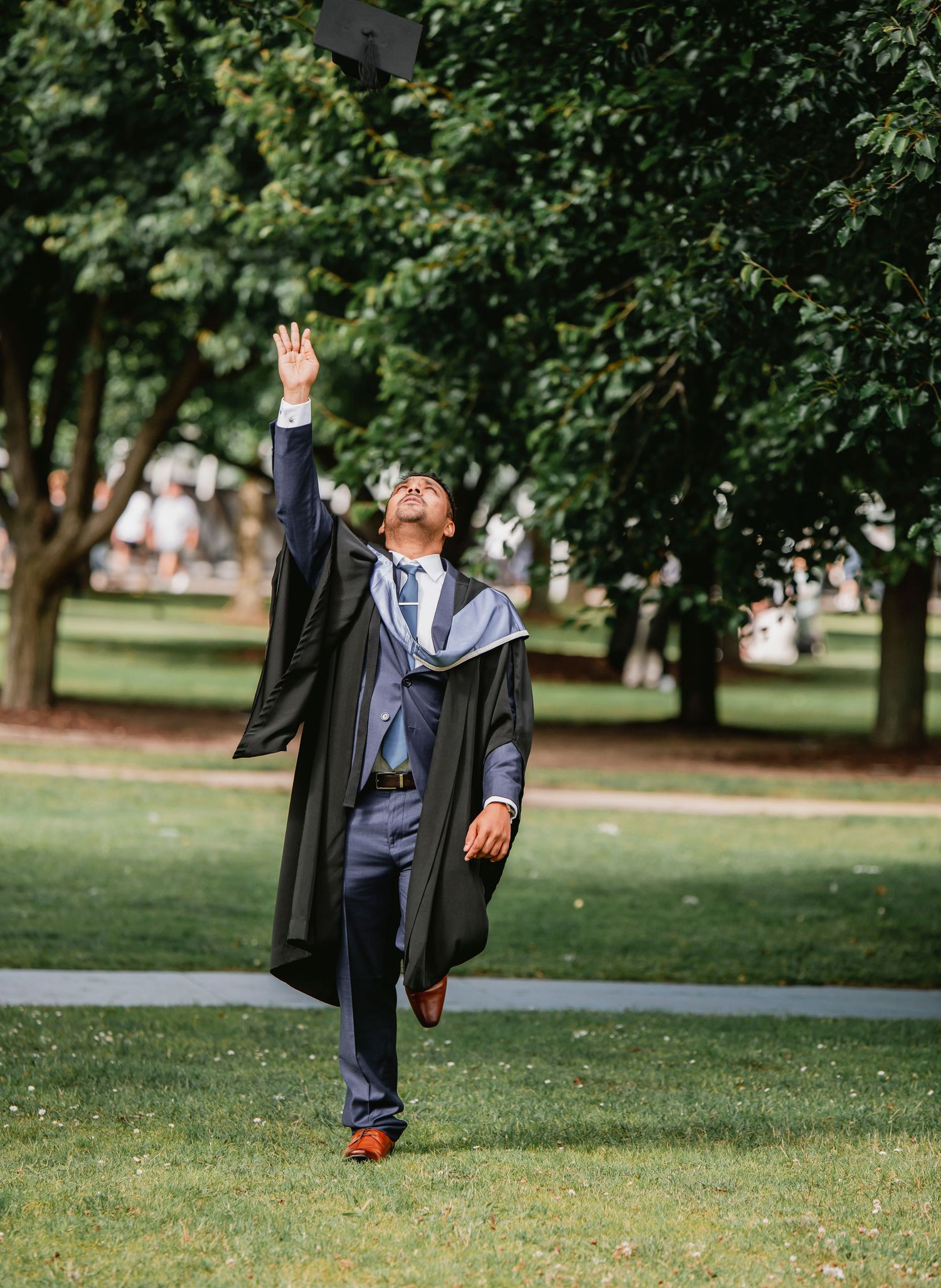 Graduate in cap and gown celebrates graduation with joyful jumps in a scenic park setting.