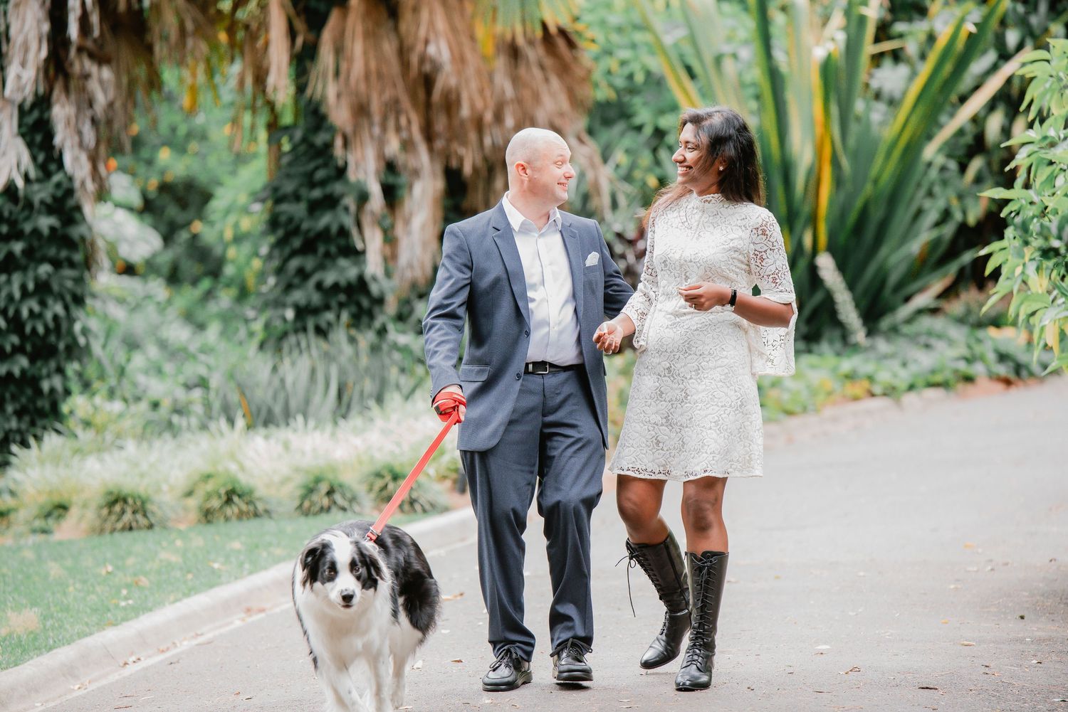 Wedding couple walks along garden path with their dog during outdoor celebration at botanical venue.