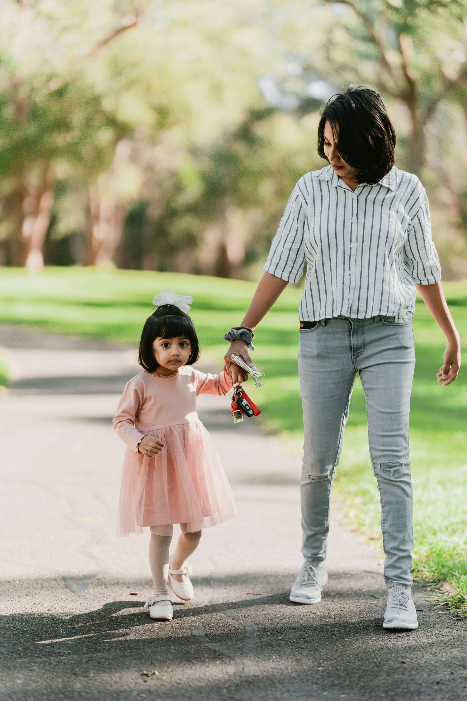 A parent and child walking together on a path through a park, with the child wearing a pink dress.