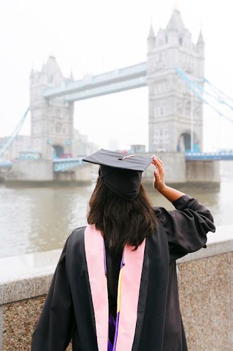 Graduate in cap and gown with pink stole looking at Tower Bridge in London on a cloudy day.