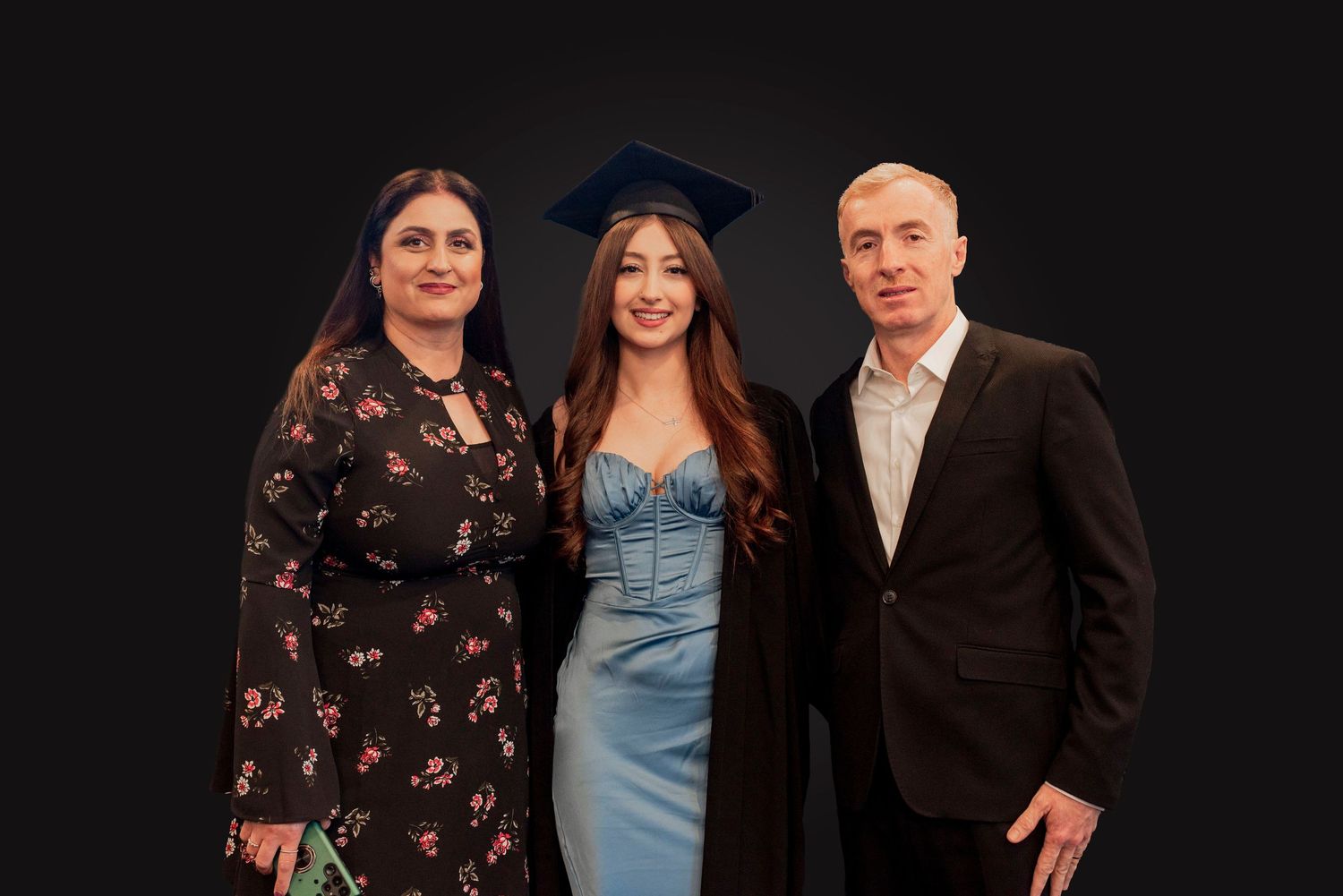 A graduate in blue dress and cap poses with two others in formal attire against a dark background.