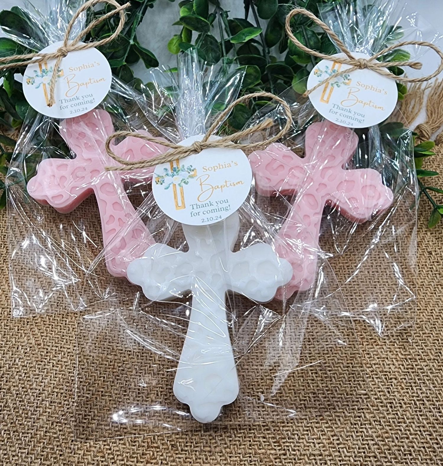 Pink and white decorative cross ornaments wrapped in cellophane with twine and gift tags on burlap background.