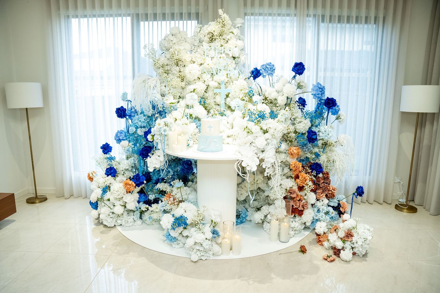 White and blue floral balloon arch installation with orange accents decorates a bright indoor space near a window.