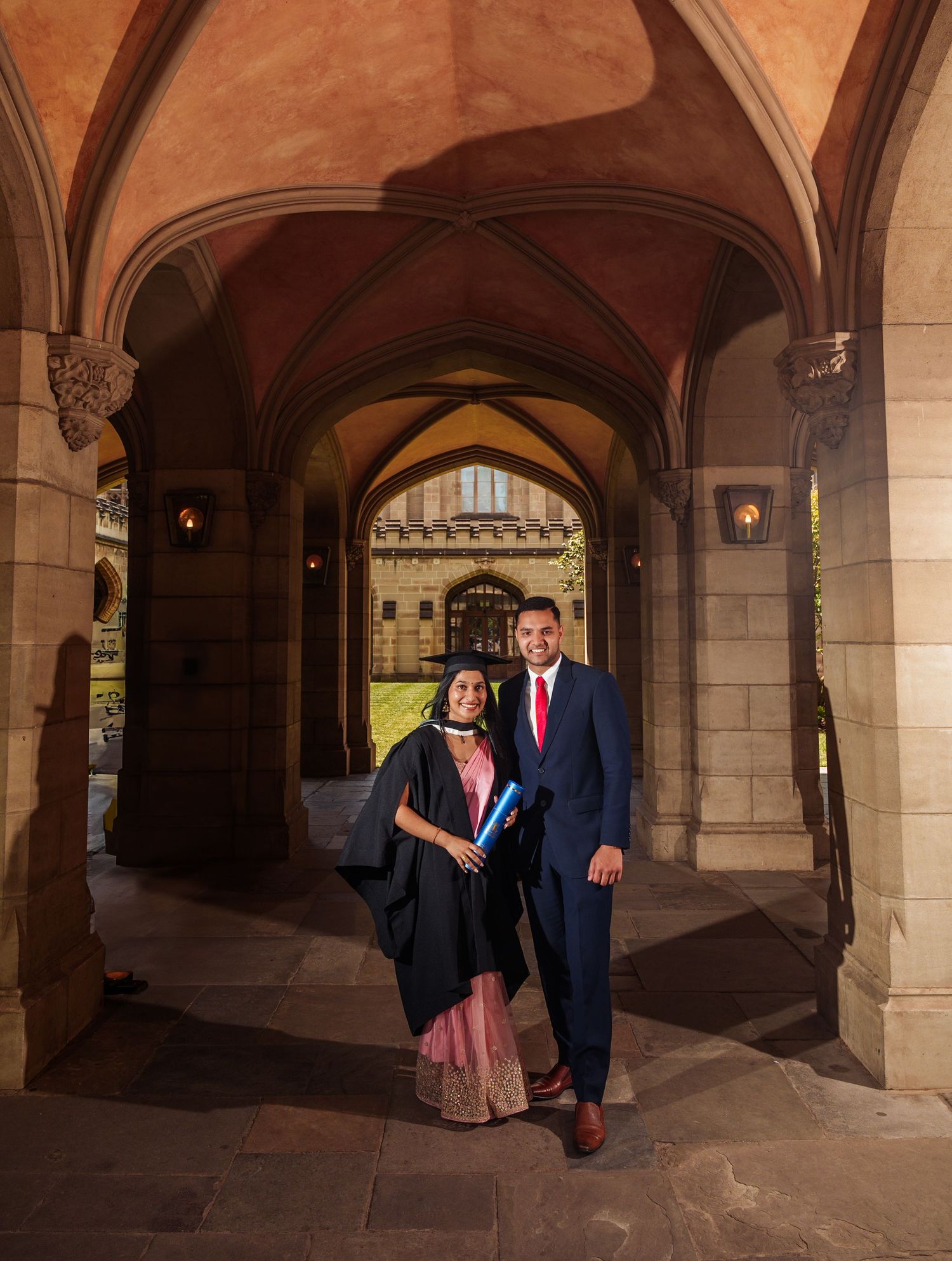 Two graduates pose in formal attire under an ornate stone archway with vaulted ceilings at a university campus.