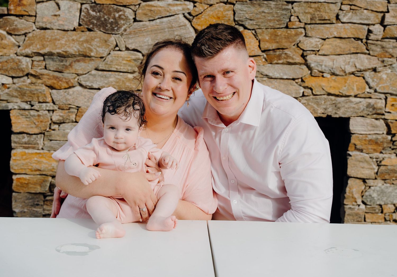Family wearing white and pink outfits poses together against rustic stone wall backdrop for portrait session.