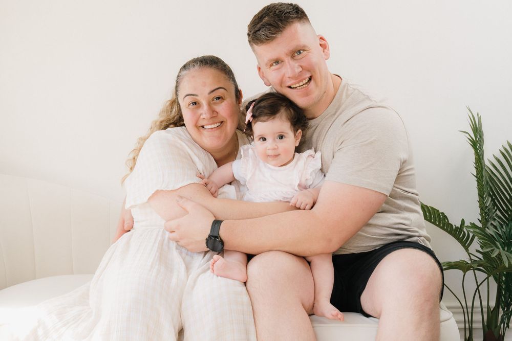 Happy family portrait with smiling parents and baby girl sitting together on white bed near green plant.