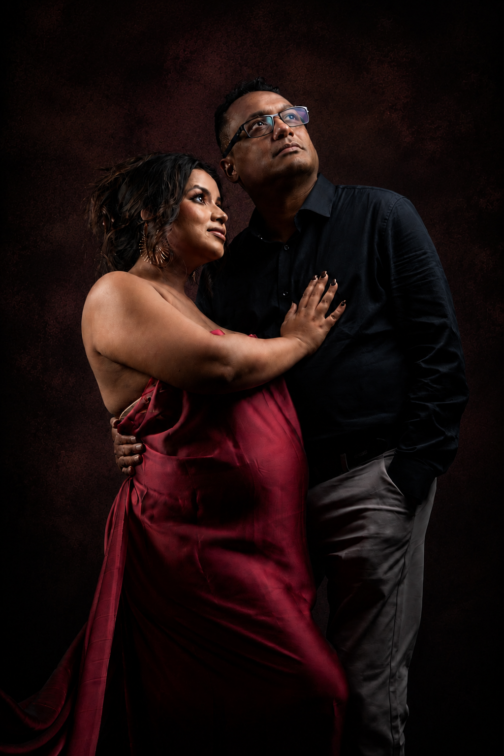 A couple poses elegantly, woman in red gown embracing man in black shirt against dark studio background.