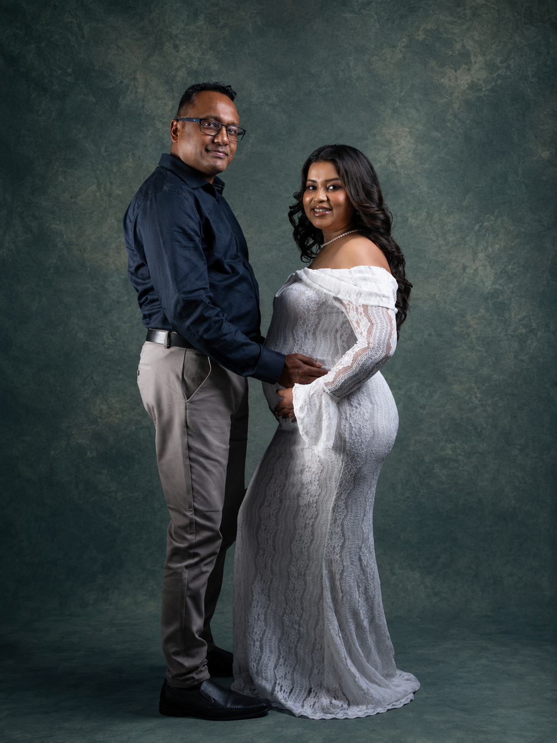 A couple poses for a maternity portrait, man in navy shirt with pregnant woman in white lace gown against gray backdrop.