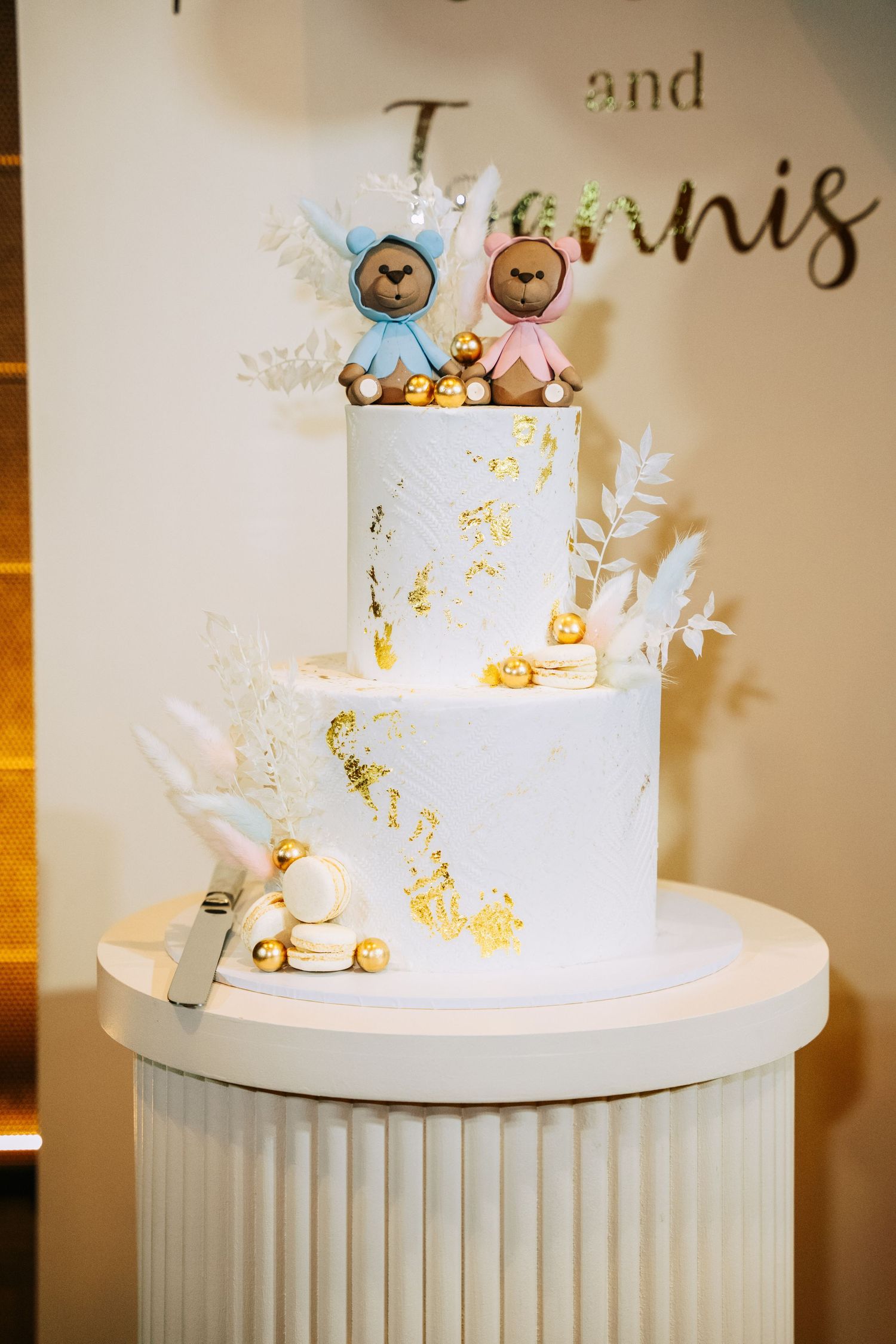 Elegant two-tier white wedding cake decorated with gold accents, delicate floral details and cute plush animal cake toppers.