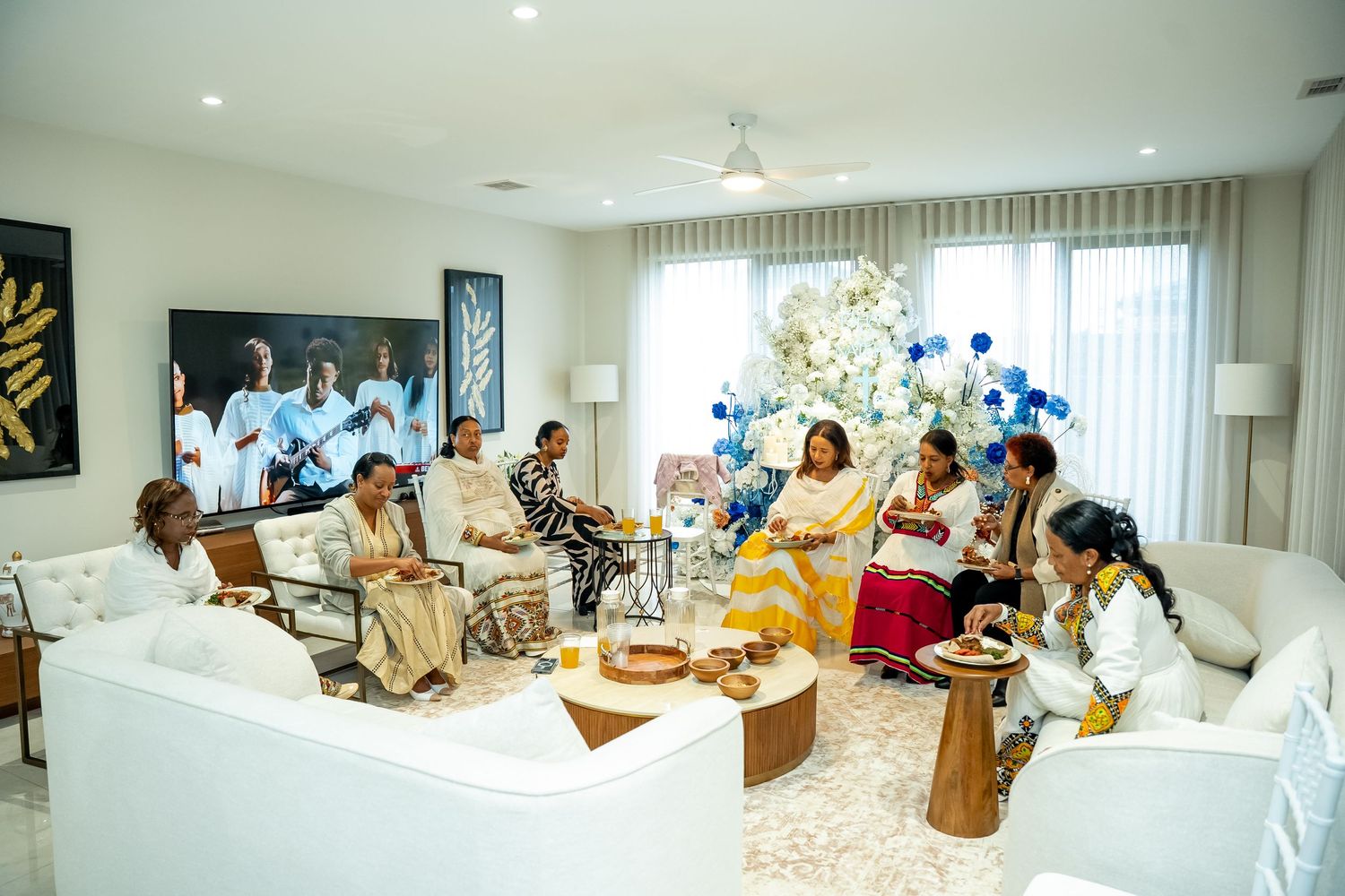 Group of people gathered in a bright white room sitting on the floor during what appears to be a social event.